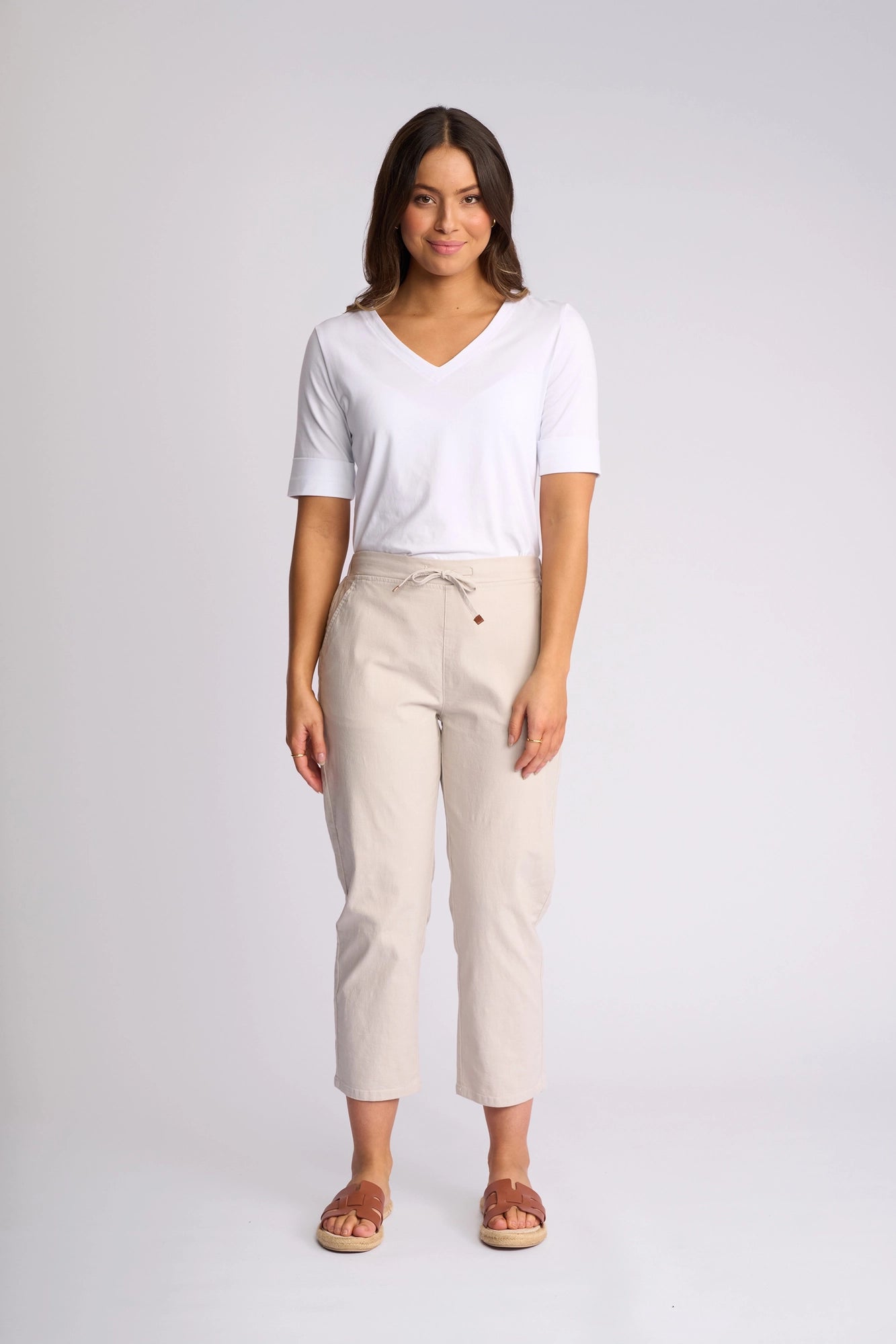 A woman stands smiling in a white short-sleeve V-neck top and Vassalli’s Henley 7/8 Pant in Stone—crafted from stretch cotton—featuring a drawstring waist and slim-leg fit. She finishes the look with brown sandals against a plain, light-colored background.