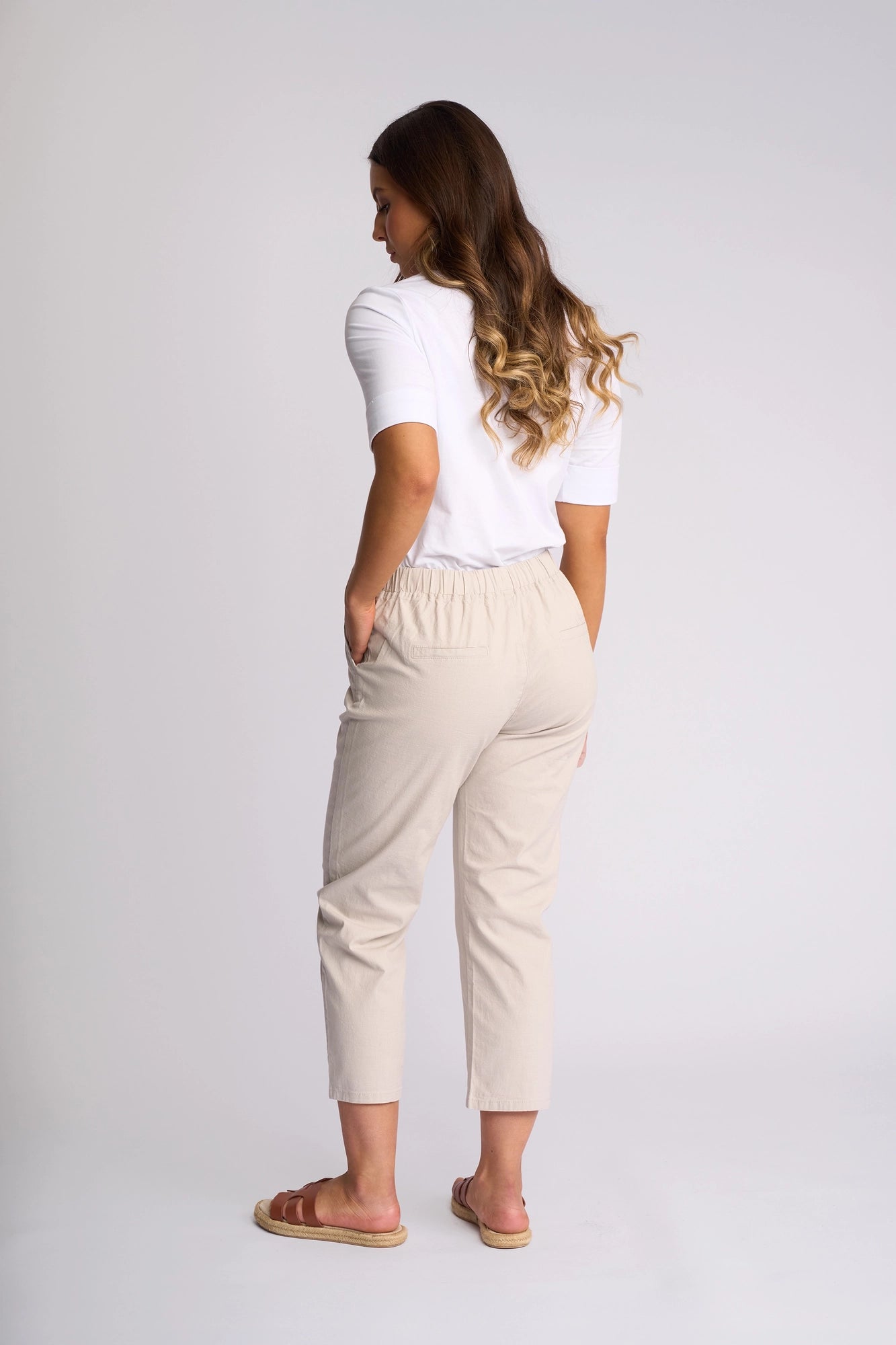 A woman with long wavy hair stands facing away, wearing a white top, Vassalli Henley 7/8 Pant in Stone, and brown sandals against a plain light background.