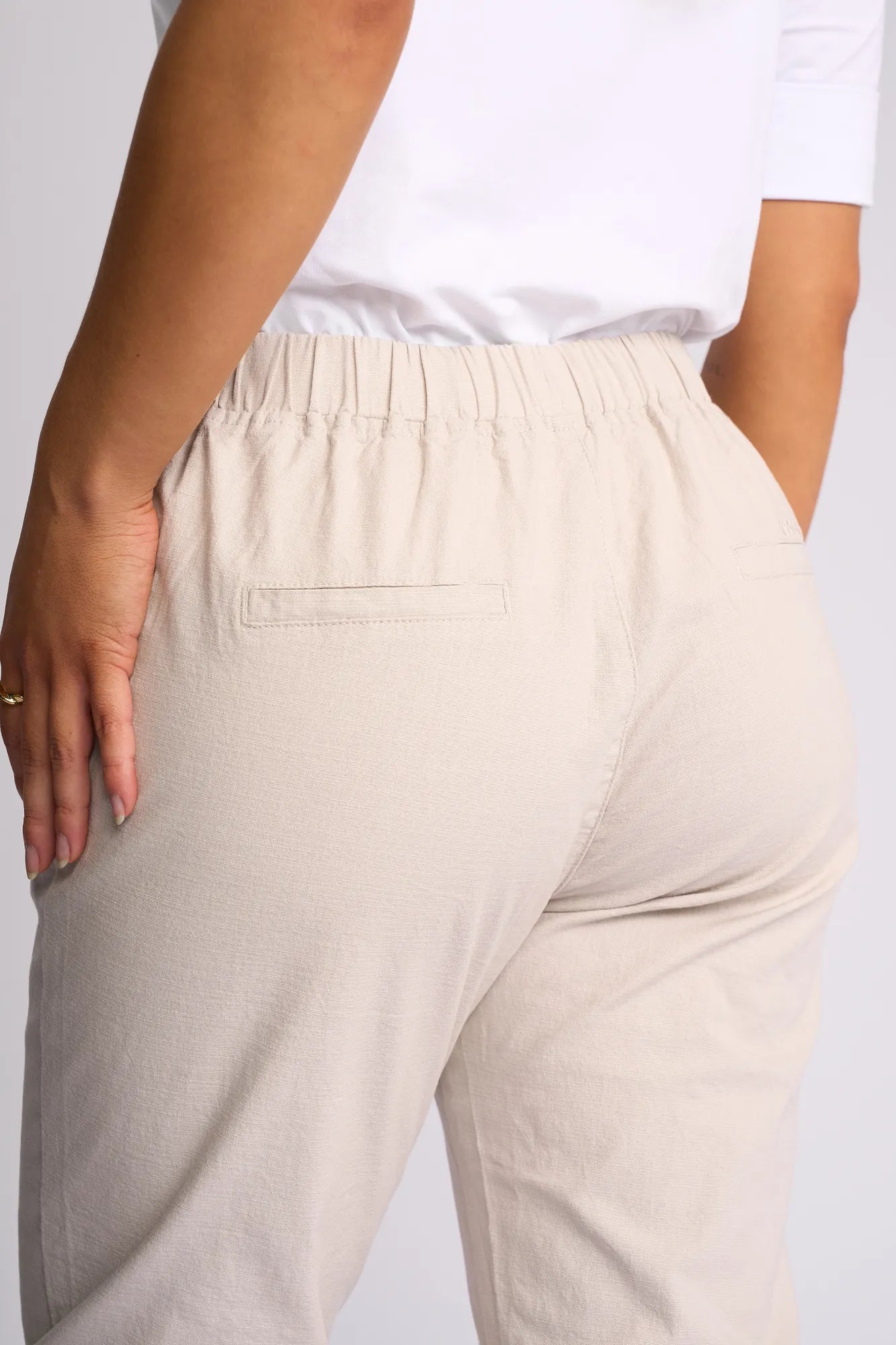 Wearing the Vassalli Henley 7/8 Pant in Stone, crafted from stretch cotton with an elastic waistband and two back welt pockets, this person pairs the light beige slim-leg trousers with a white shirt. Their left hand rests on their hip, accentuating a gold ring.