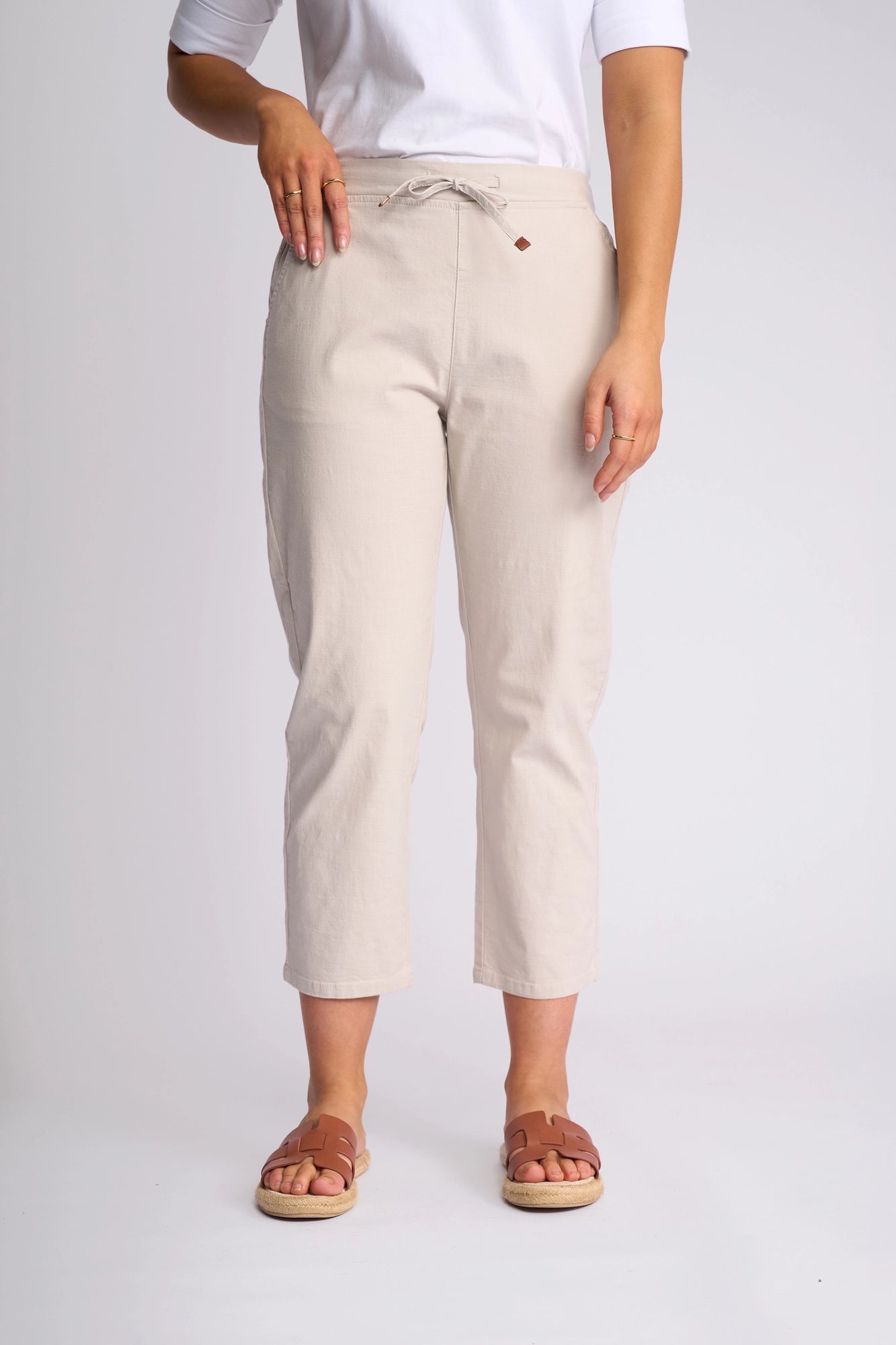 A person wearing the Vassalli Henley 7/8 Pant in Stone, styled with a white elbow-sleeve top and brown sandals, stands against a plain light background with only the lower half of the body visible.