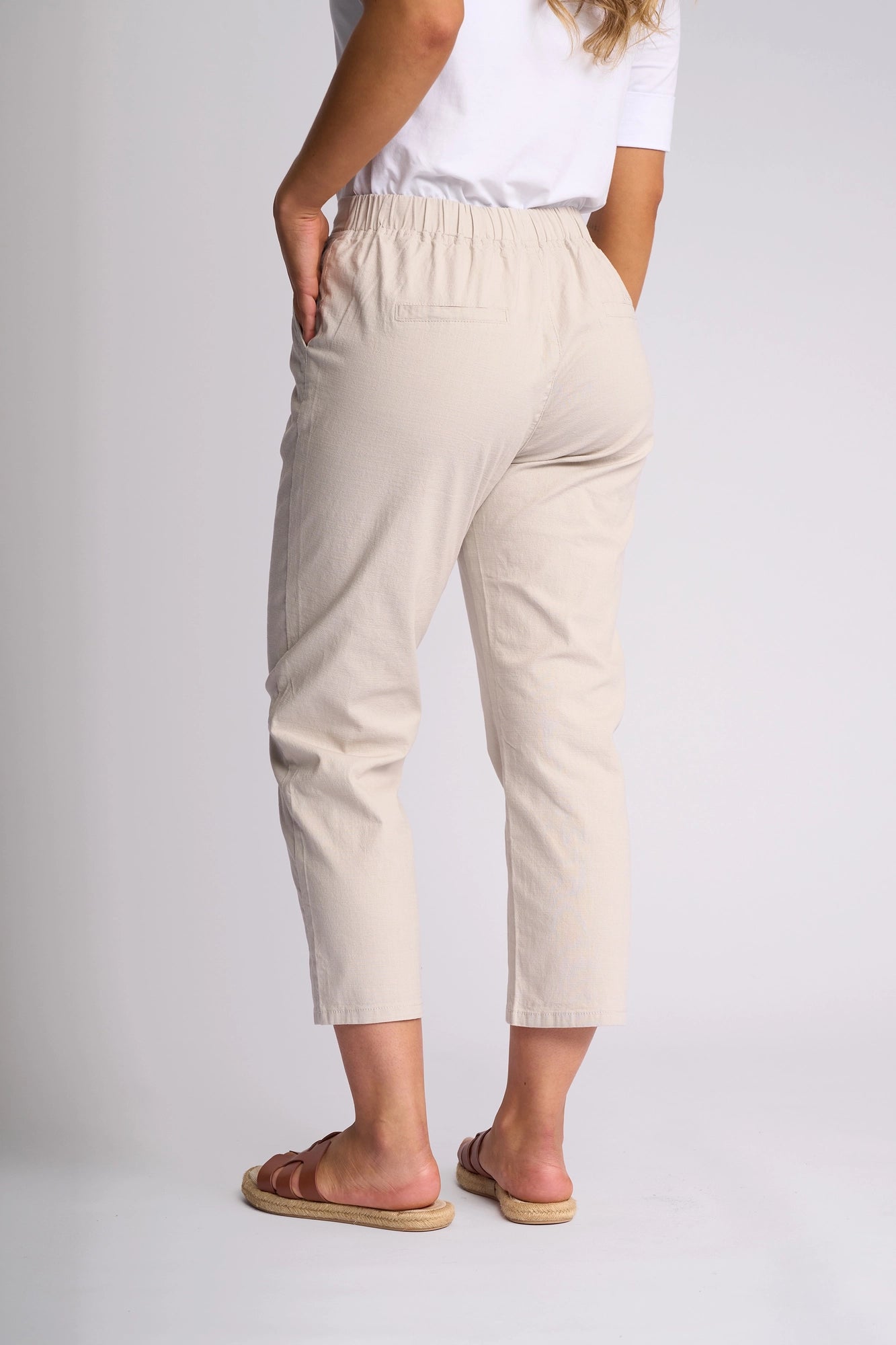 A woman stands facing away, hands in pockets, wearing the Vassalli Henley 7/8 Pant in Stone, paired with a white shirt and brown sandals against a plain light background.