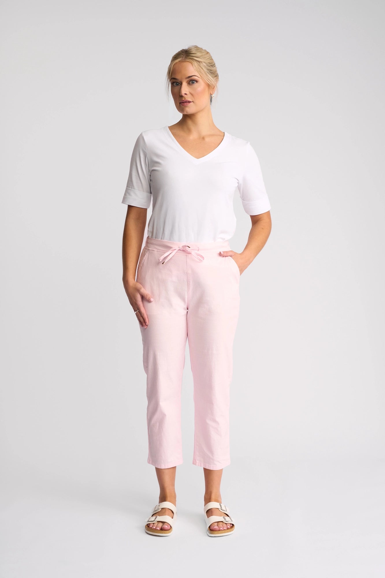 A woman stands against a plain background wearing a white V-neck shirt, the Vassalli Henley 7/8 Pant - Taffy in stretch cotton with a waist tie and slim-leg silhouette, and white sandals. She has blonde hair tied back and one hand in her pocket.