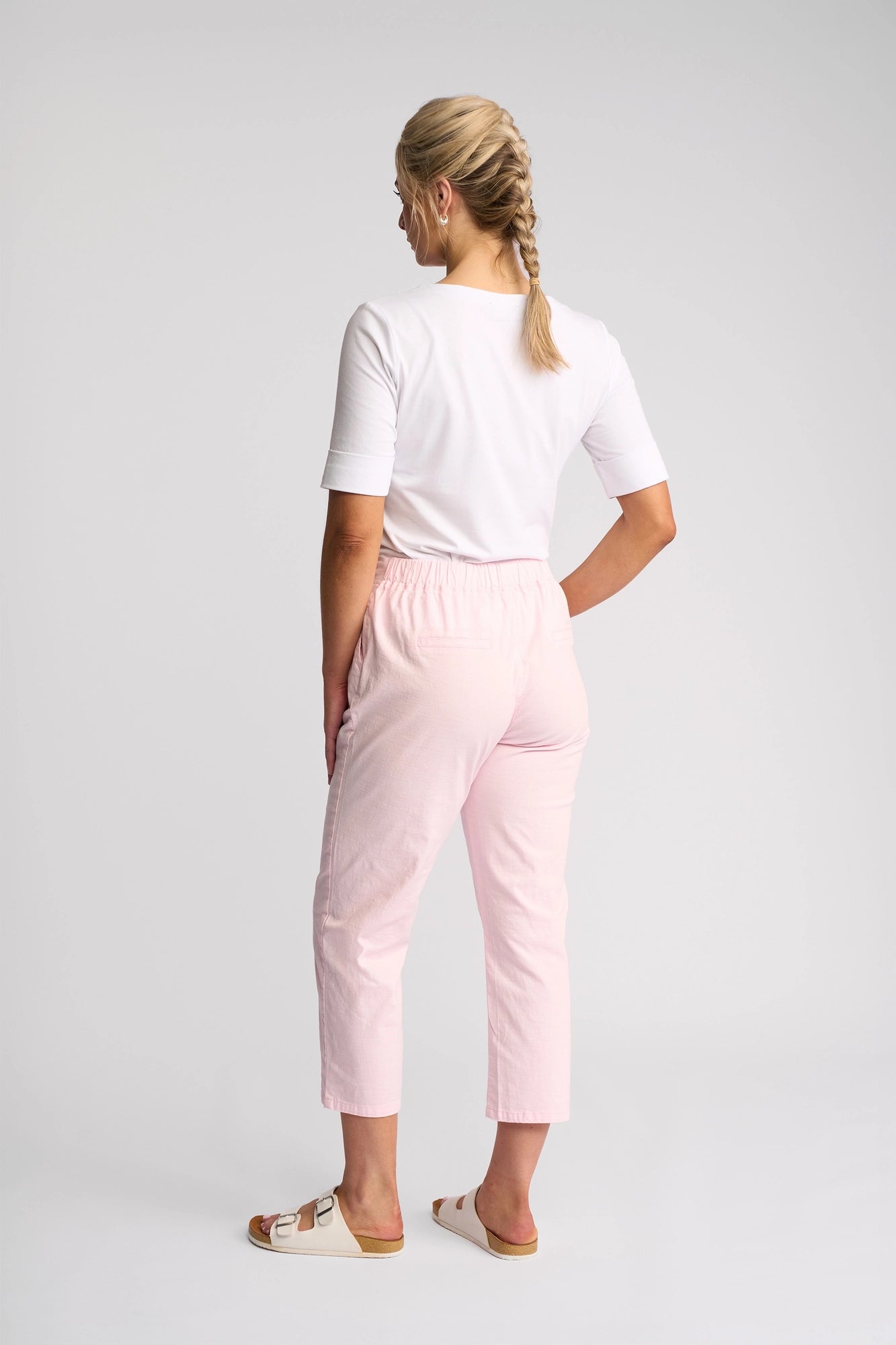 A woman with blond braided hair faces away from the camera, wearing a white shirt, the Vassalli Henley 7/8 Pant in Taffy, and white sandals, standing against a plain light background.