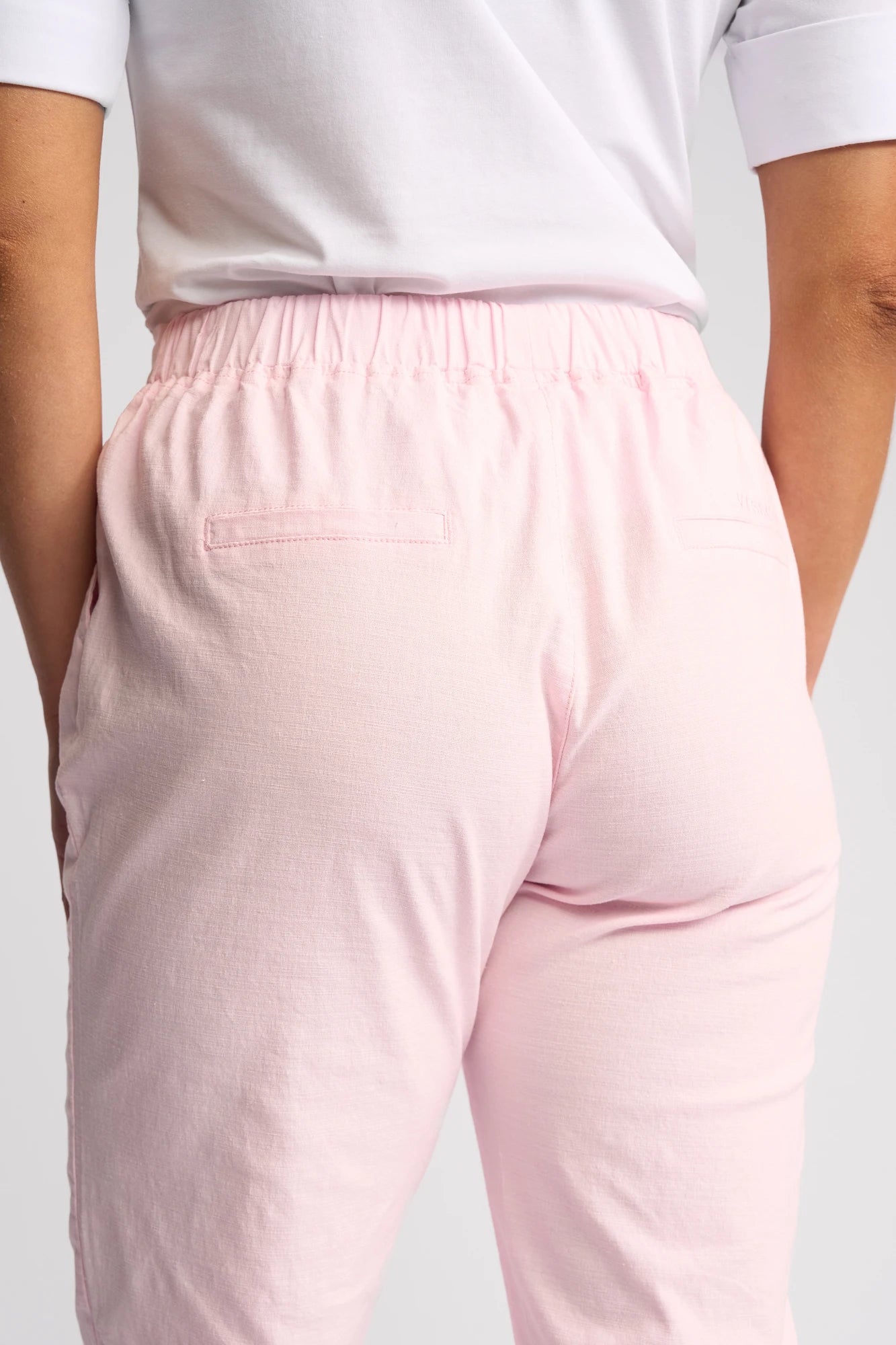 A person is shown from the back with hands in pockets, wearing a white shirt and Vassalli’s Henley 7/8 Pant in Taffy—a light pink stretch cotton slim-leg pant featuring an elastic waistband.