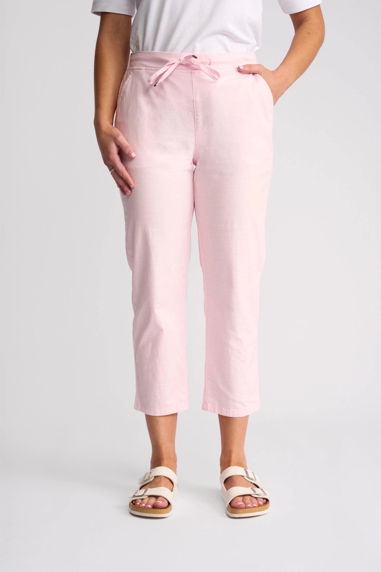 A person wearing Vassalli’s Henley 7/8 Pant in Taffy, paired with a white top and white sandals, stands with one hand in their pocket against a plain background.
