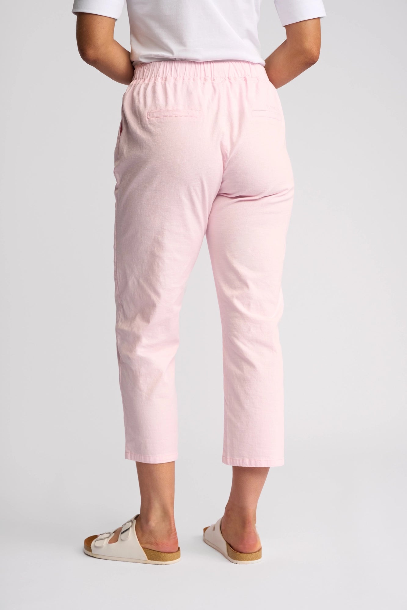 A person is shown from the back wearing Vassalli’s Henley 7/8 Pant in Taffy—a light pink, slim-leg style made from comfortable stretch cotton with an elastic waistband and two back pockets. They pair the pants with white sandals and a white shirt, set against a plain, light-colored background.