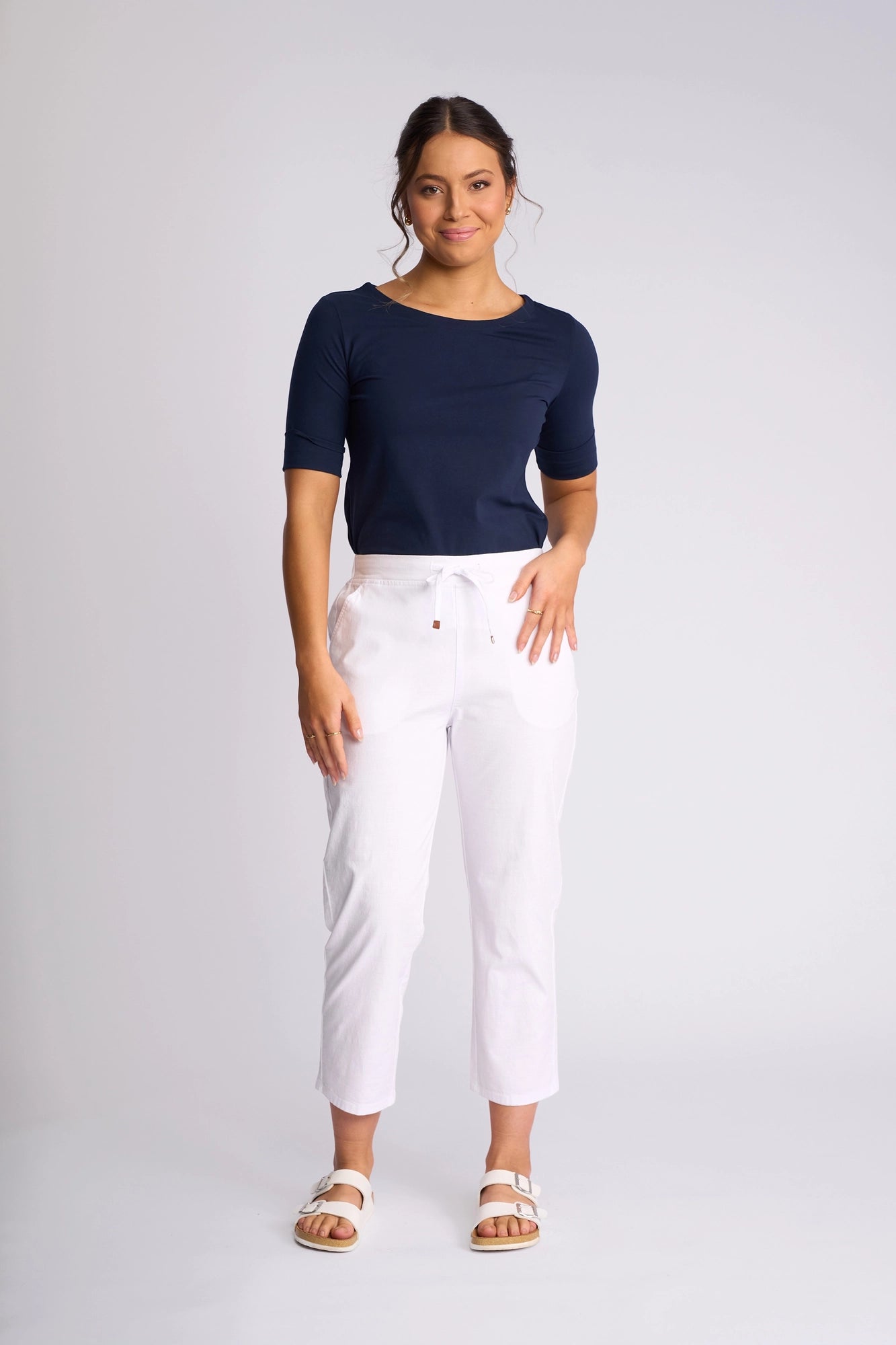 A woman stands against a plain background, wearing a navy blue top and the Vassalli Henley 7/8 Pant in white, made from stretch cotton with a drawstring waist. She pairs the look with white sandals, her dark hair tied back and a relaxed, confident expression.