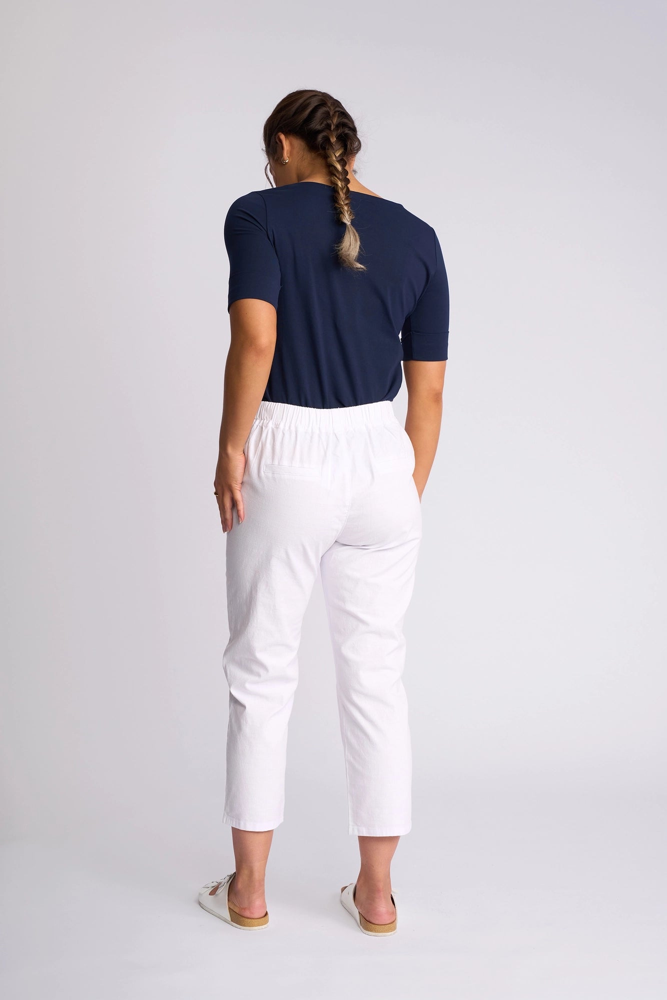 A woman with a braid, dressed in a navy top and Vassalli Henley 7/8 Pant - White, stands facing away from the camera against a plain light background.