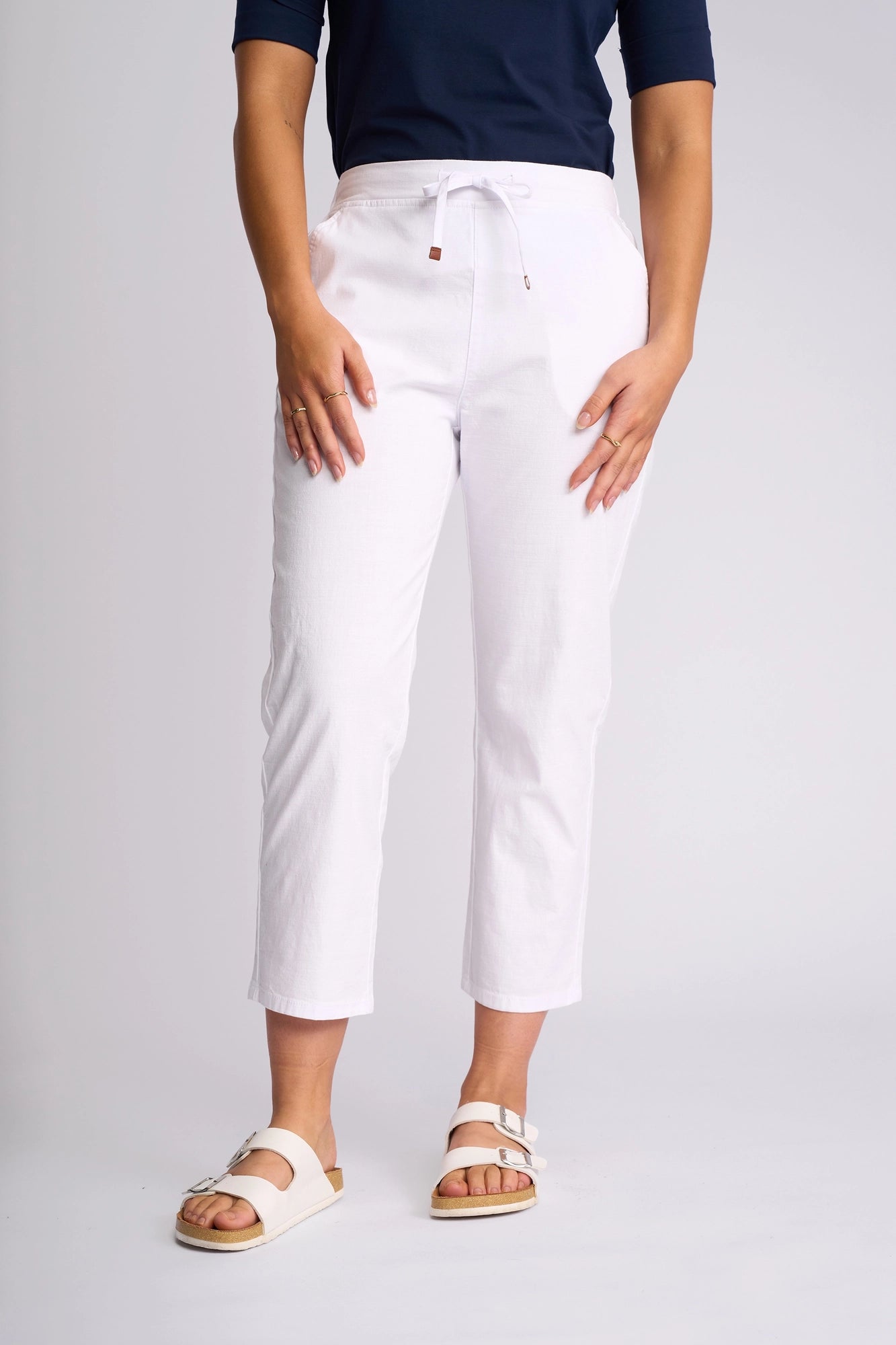 A person wearing a navy top, Vassalli Henley 7/8 Pant in white made from stretch cotton, and white sandals stands against a plain light background with hands resting on their thighs.
