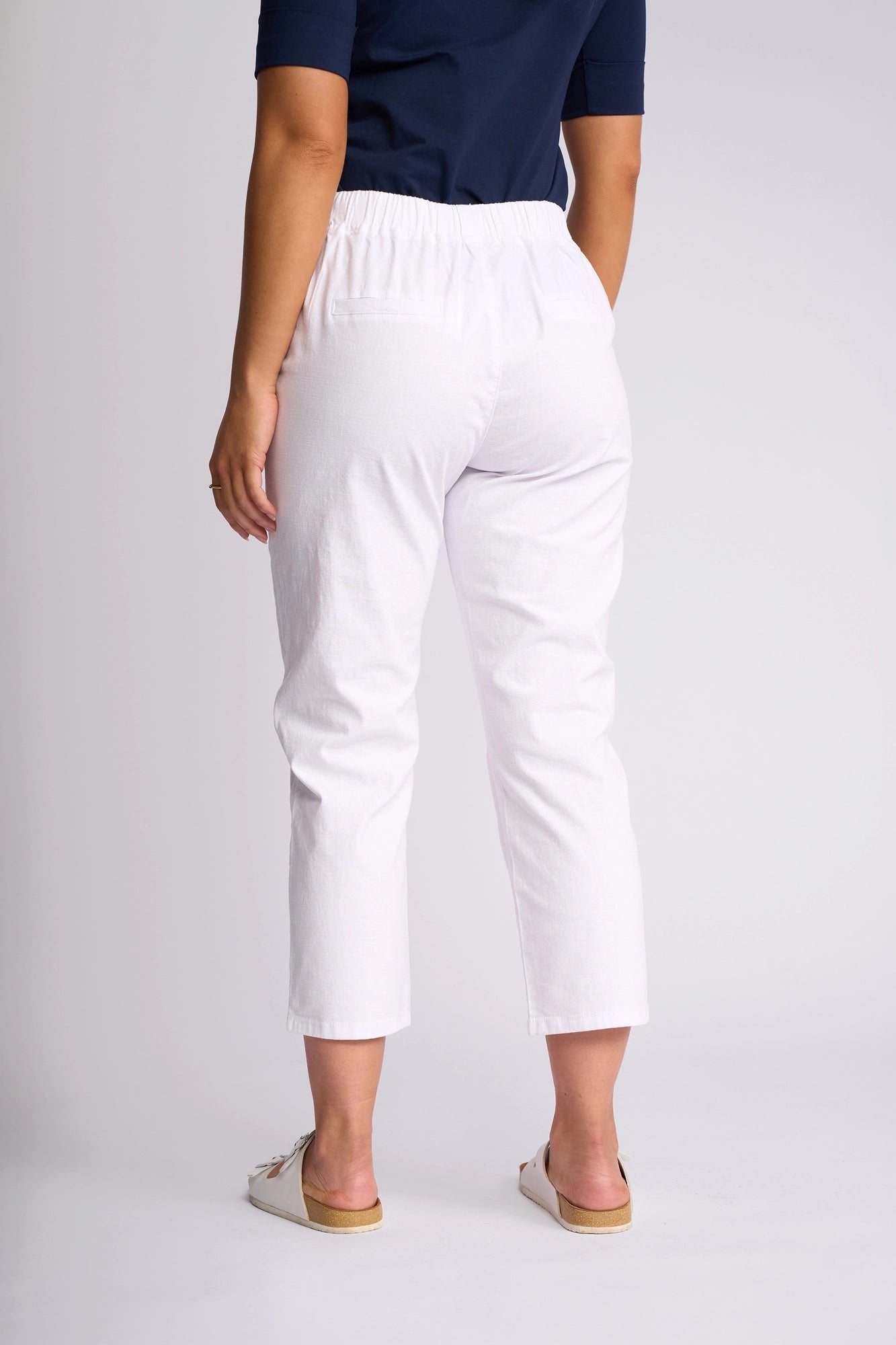 A person wearing a navy top, Vassalli Henley 7/8 Pant in white with an elastic waistband, and white slip-on shoes stands facing away against a plain background.
