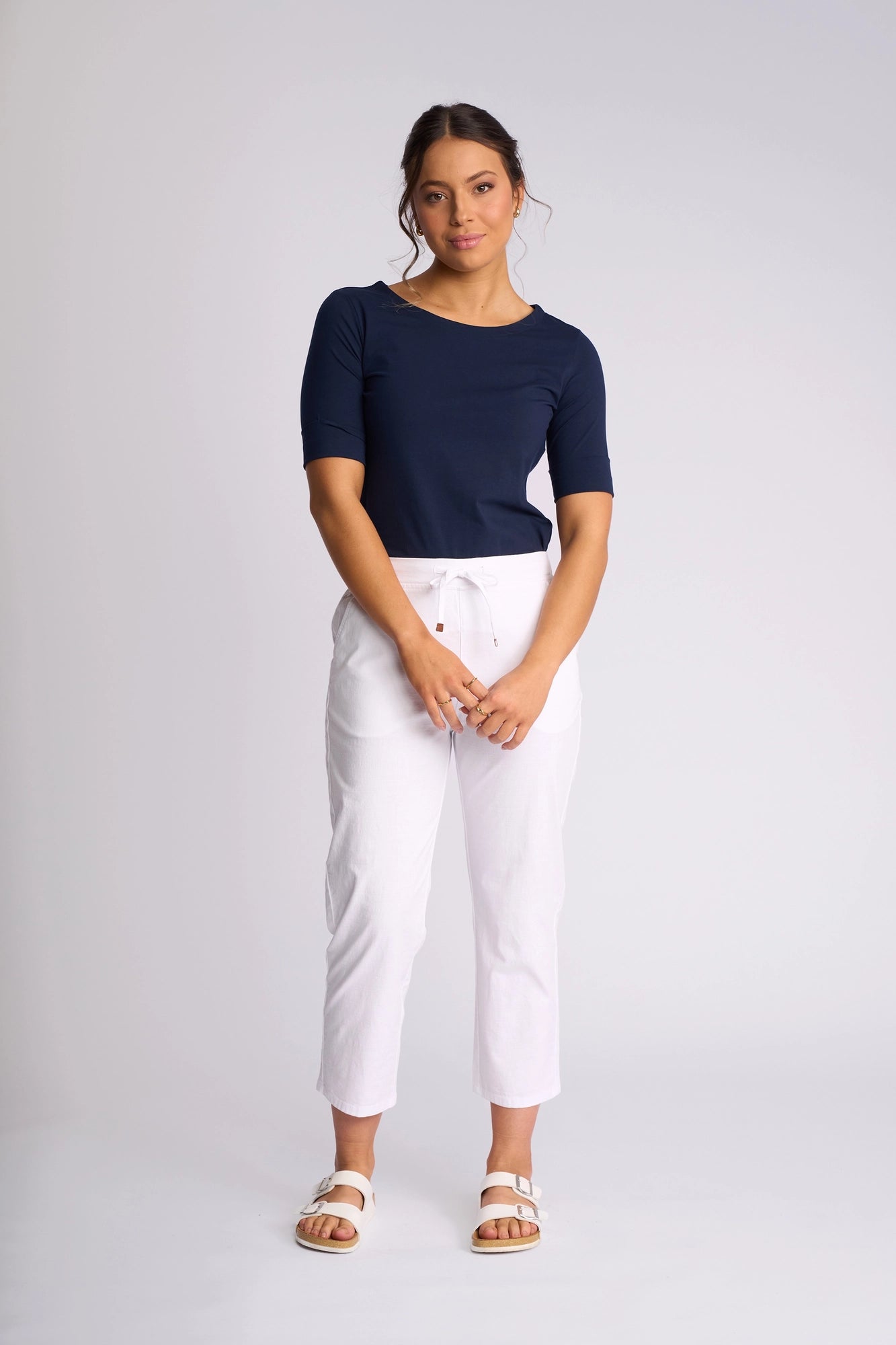 A woman stands against a plain background, wearing a navy-blue top, Vassalli Henley 7/8 Pant in white with a slim-leg silhouette, and white sandals, her hands loosely clasped in front of her.