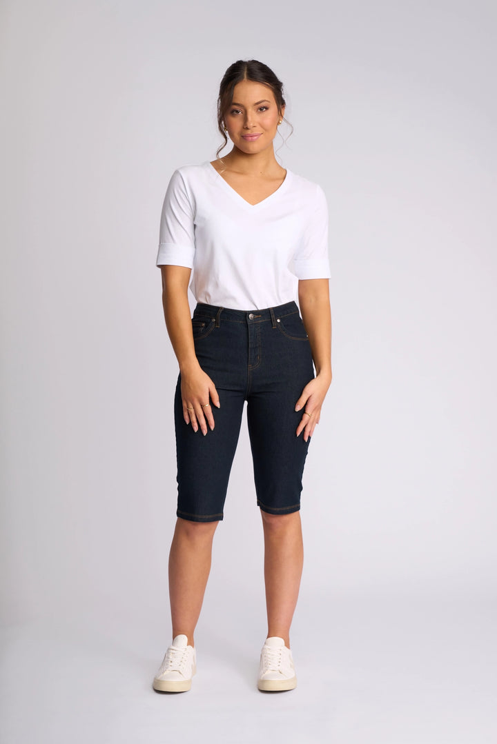 A woman stands facing forward against a plain light gray background, wearing a white short-sleeve V-neck shirt, the Vassalli Alfa Short - Indigo Denim (Contrast Stitch) crafted from lightweight stretch denim and falling to the knee, and white sneakers.