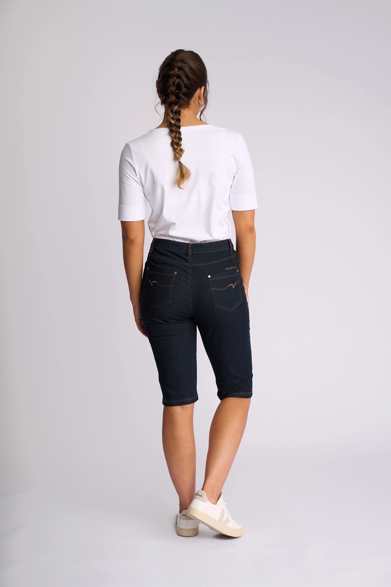 A person with a braid stands facing away, wearing a white t-shirt, white sneakers, and Vassalli’s Alfa Short in Indigo Denim (Contrast Stitch), which features a slim-leg fit, against a plain light background.
