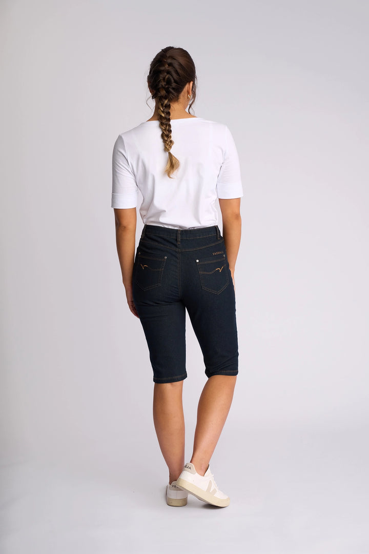 A person with a braid stands facing away, wearing a white t-shirt, white sneakers, and Vassalli’s Alfa Short in Indigo Denim (Contrast Stitch), which features a slim-leg fit, against a plain light background.
