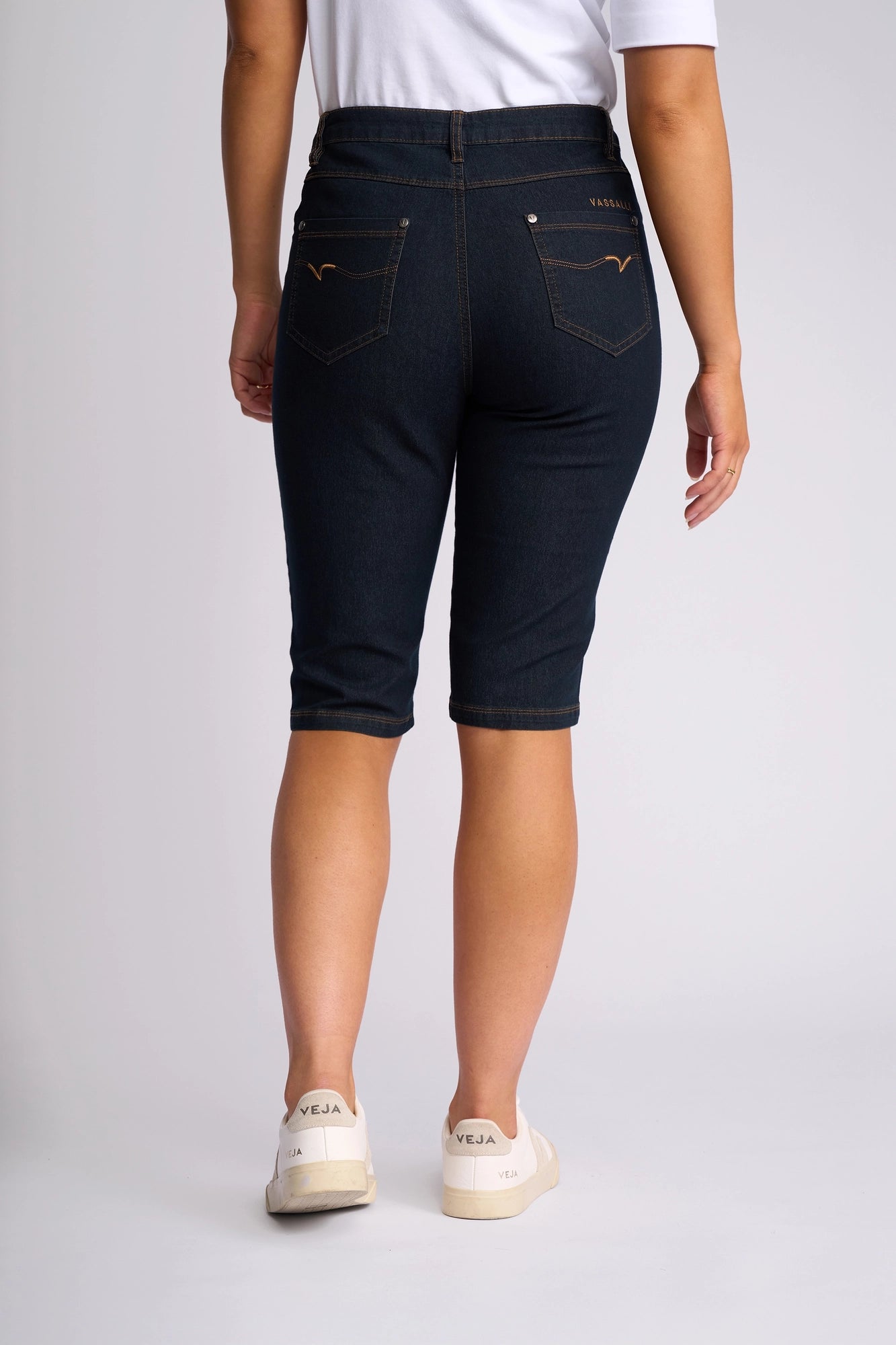 A person wearing a white shirt, Vassalli Alfa Short - Indigo Denim (Contrast Stitch) slim-leg knee-length shorts, and white Veja sneakers stands facing away against a plain light background.
