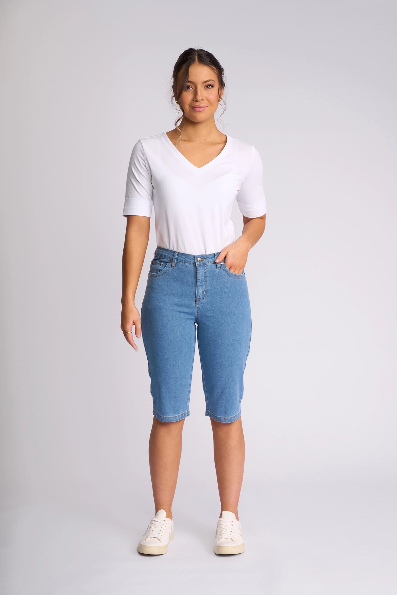 A woman stands facing forward wearing a white V-neck shirt, Vassalli’s Alfa Short - Light Denim (Contrast Stitch) slim-leg denim shorts crafted from lightweight stretch denim, and white sneakers against a plain light background—an effortless summer staple.