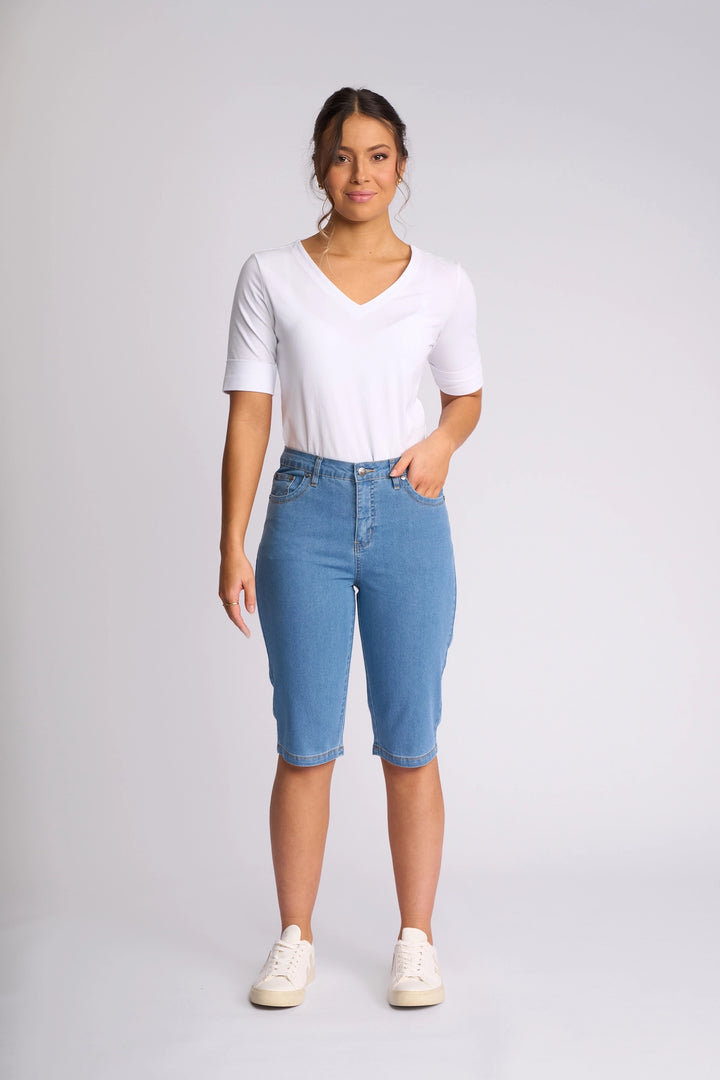 A woman stands facing forward wearing a white V-neck shirt, Vassalli’s Alfa Short - Light Denim (Contrast Stitch) slim-leg denim shorts crafted from lightweight stretch denim, and white sneakers against a plain light background—an effortless summer staple.