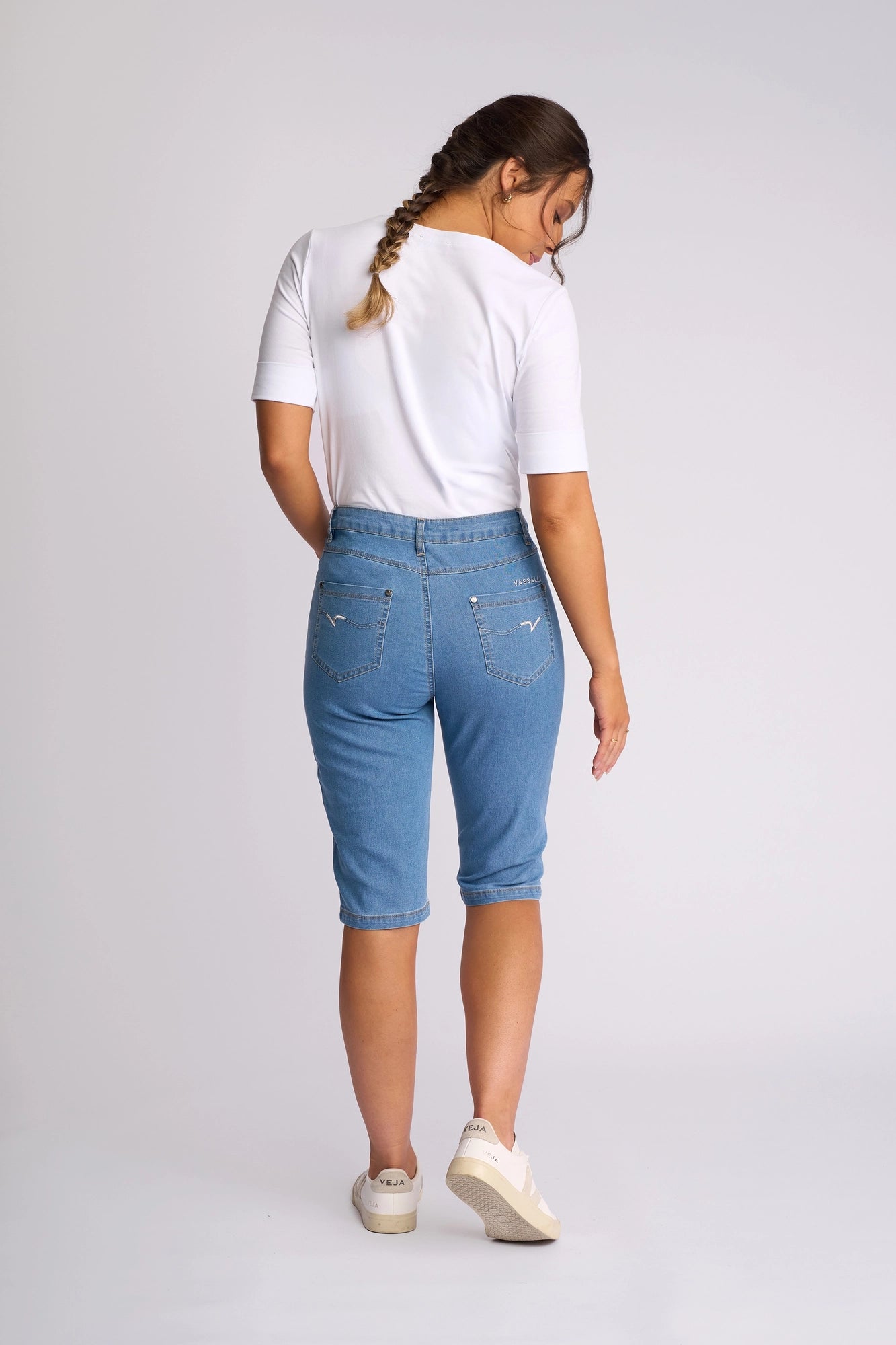 A woman with a braid, dressed in a white shirt, the Vassalli Alfa Short - Light Denim (Contrast Stitch)—a blue lightweight stretch denim knee-length short that’s perfect for summer—and beige sneakers, stands facing away against a plain white background.
