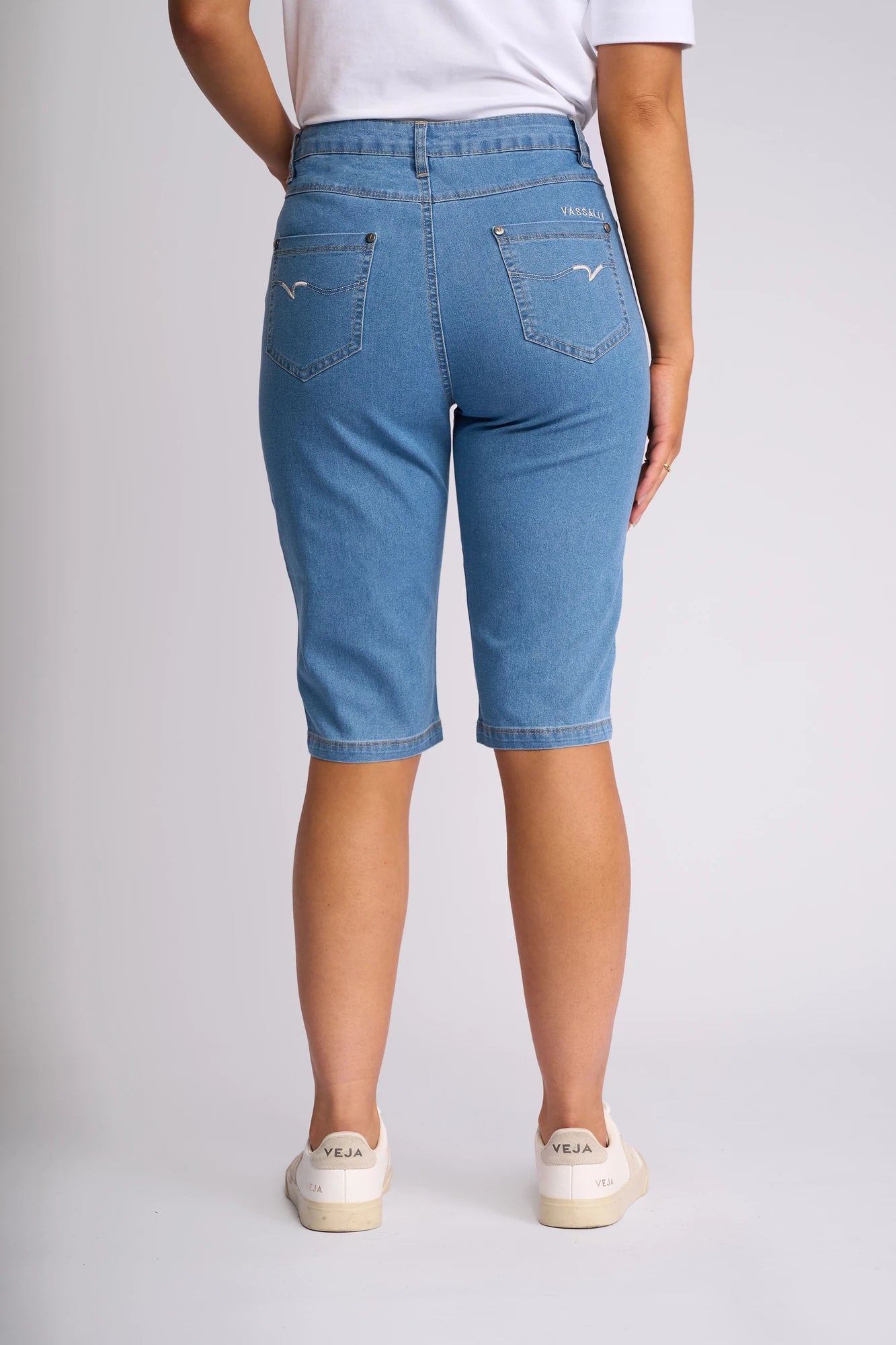 A person stands with their back to the camera, wearing Vassalli’s Alfa Short - Light Denim (Contrast Stitch), slim-leg Bermuda shorts crafted from lightweight stretch denim. The shorts are paired with white Veja sneakers, and the top of a white shirt is partially visible. The background is plain and light-colored.