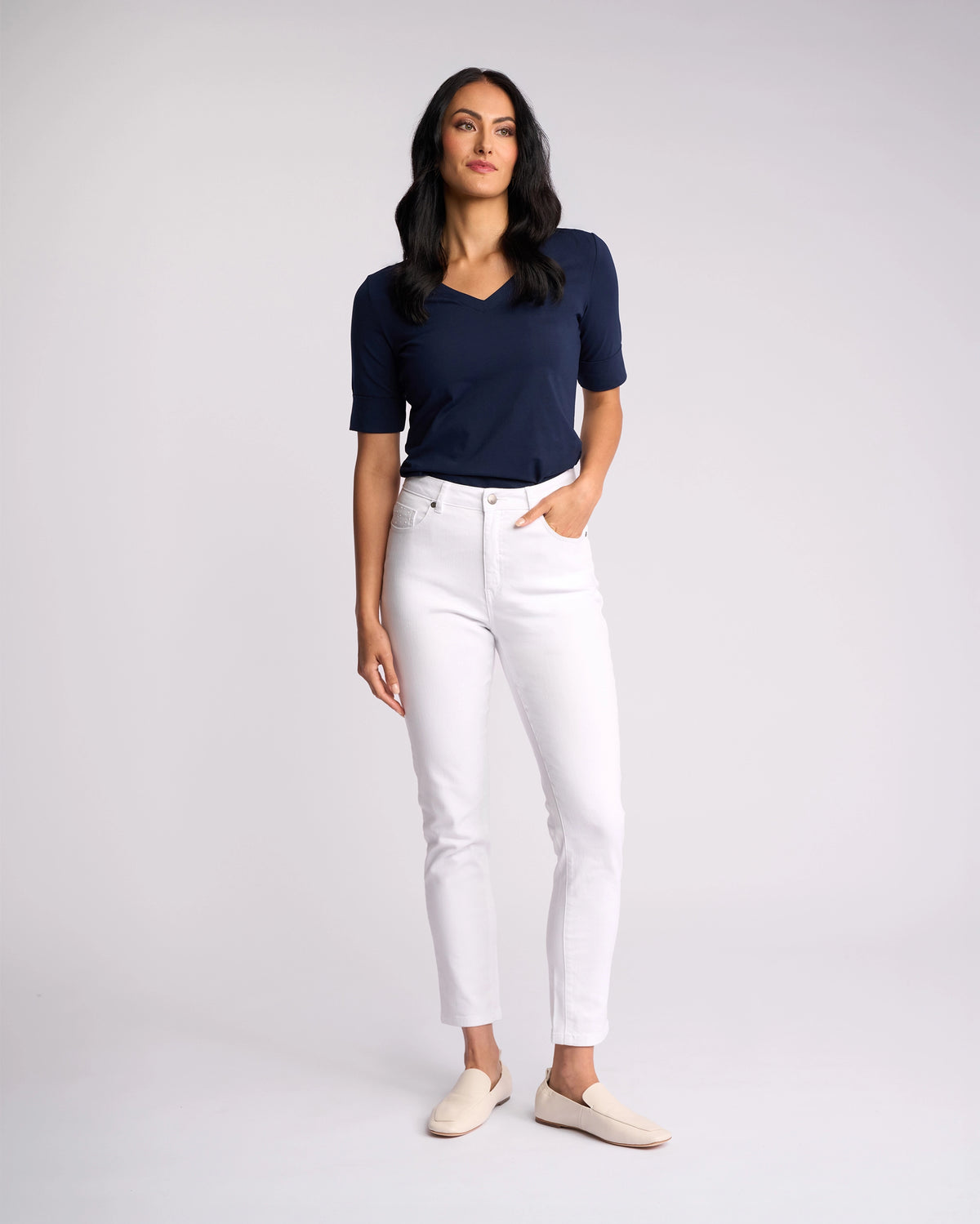 A woman stands against a plain background, wearing Vassalli’s Tide Jean in white denim, paired with a navy short-sleeve top and beige slip-on shoes. She has one hand in her pocket and gazes slightly to the side.