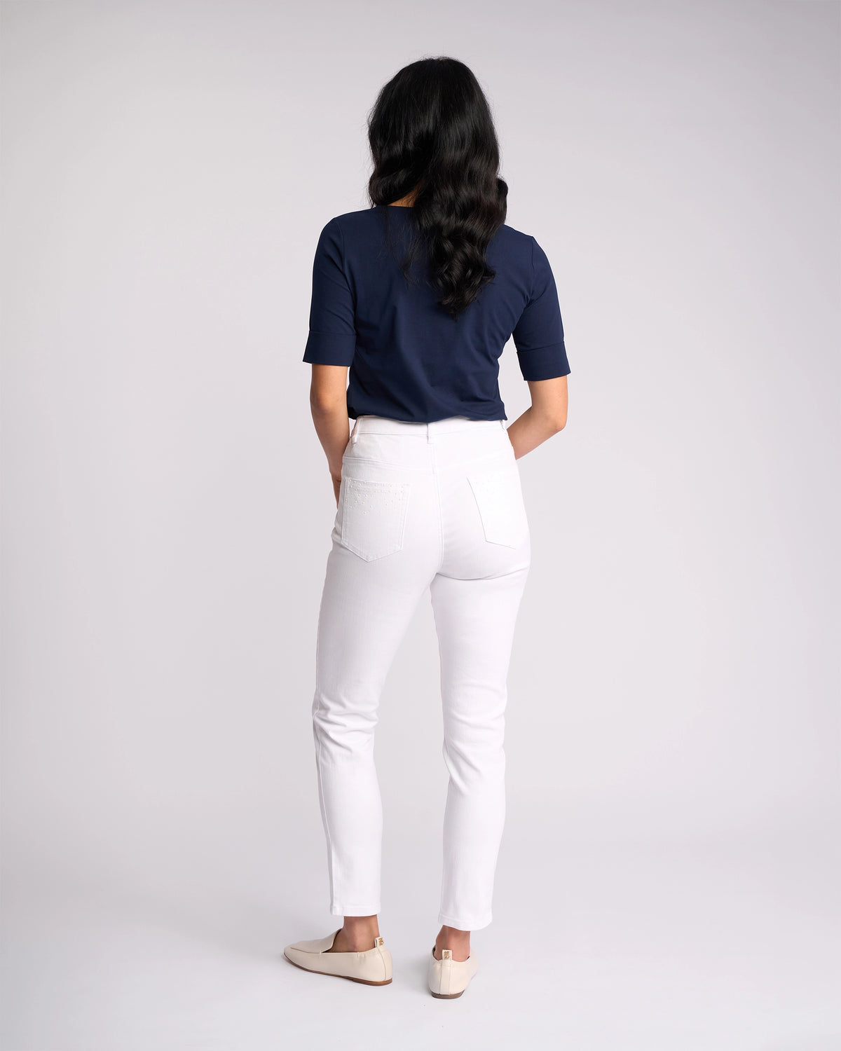 A person with long, wavy dark hair stands facing away, wearing a navy short-sleeve top, Vassalli Tide Jean - White Denim, and cream shoes against a plain light background.