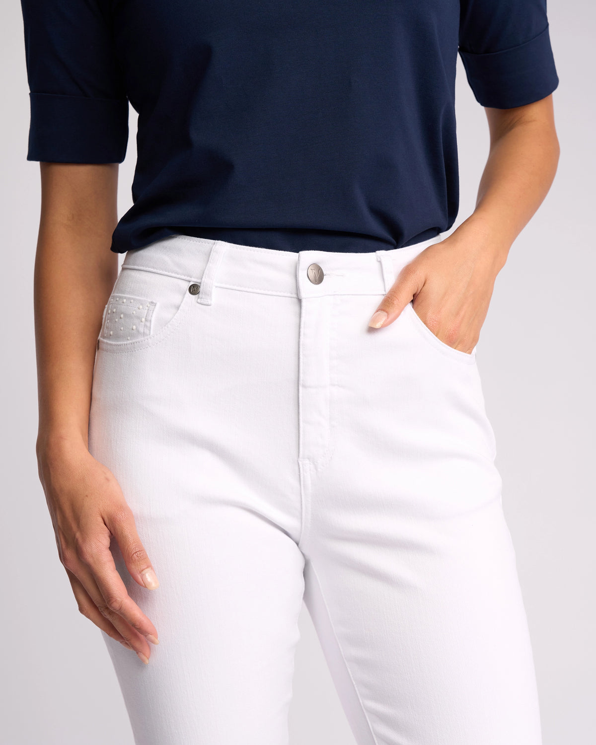 A person models the Vassalli Tide Jean in white denim, paired with a navy blue short-sleeve top, shown from shoulders to thighs against a plain background, with one hand tucked into the jeans' pocket.