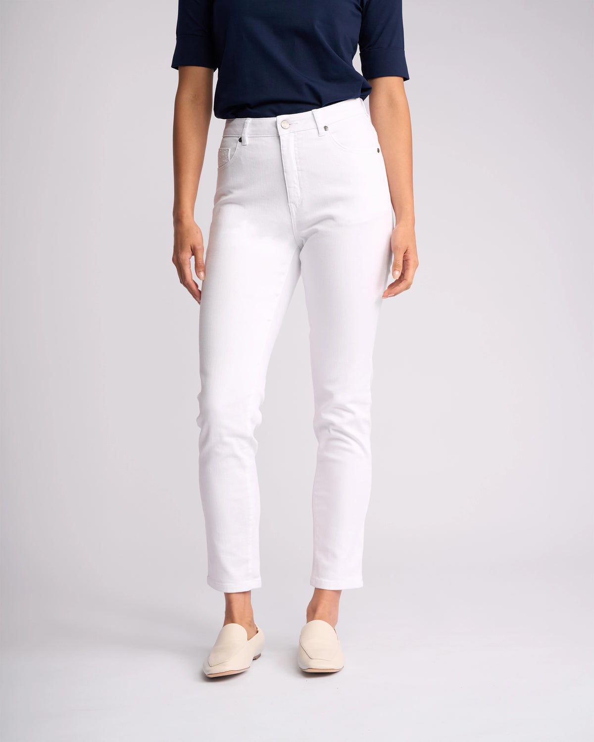 Wearing a navy short-sleeve top, Vassalli's Tide Jean in White Denim, and cream slip-on shoes, the headless individual poses against a plain light background.