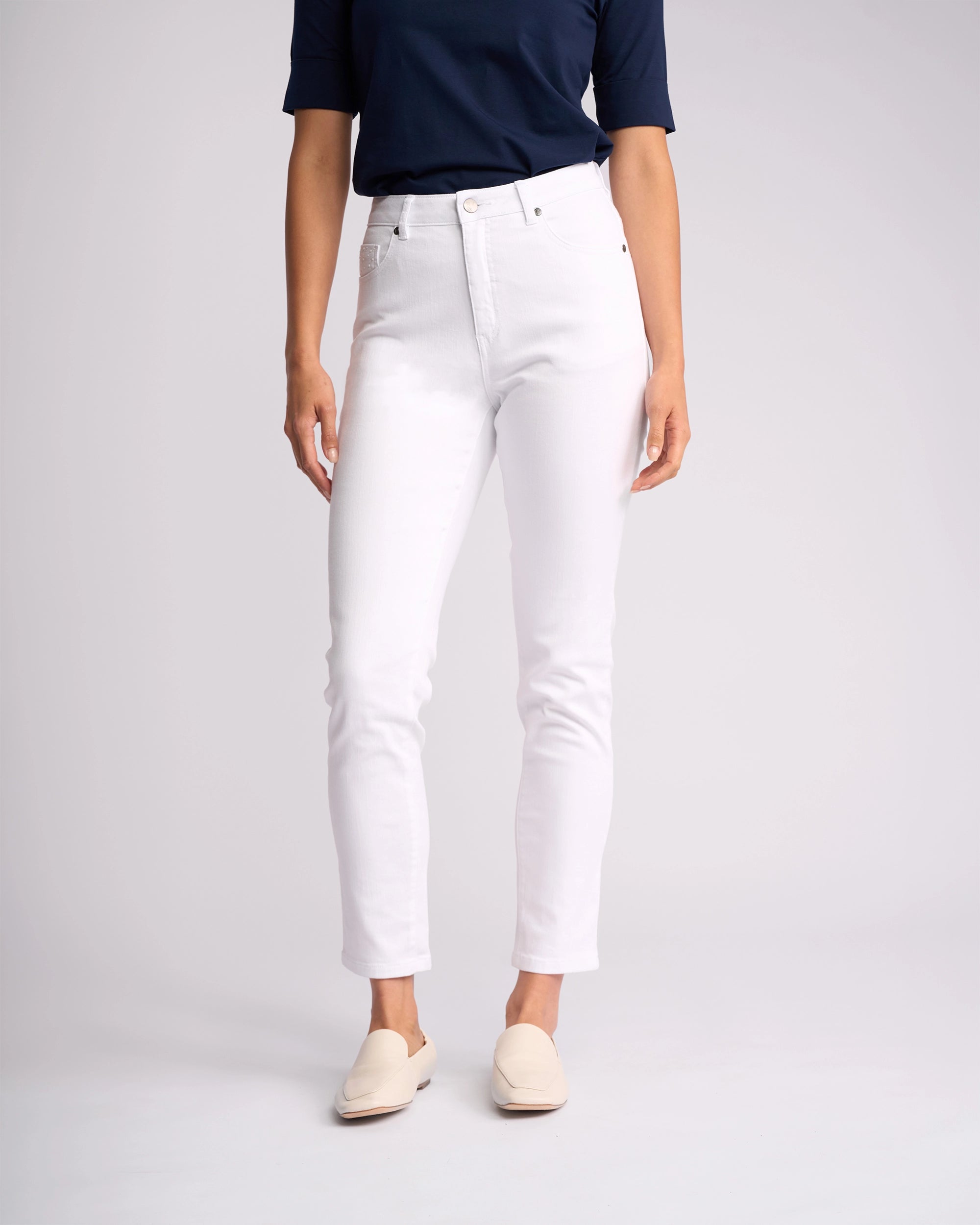 Wearing a navy short-sleeve top, Vassalli's Tide Jean in White Denim, and cream slip-on shoes, the headless individual poses against a plain light background.