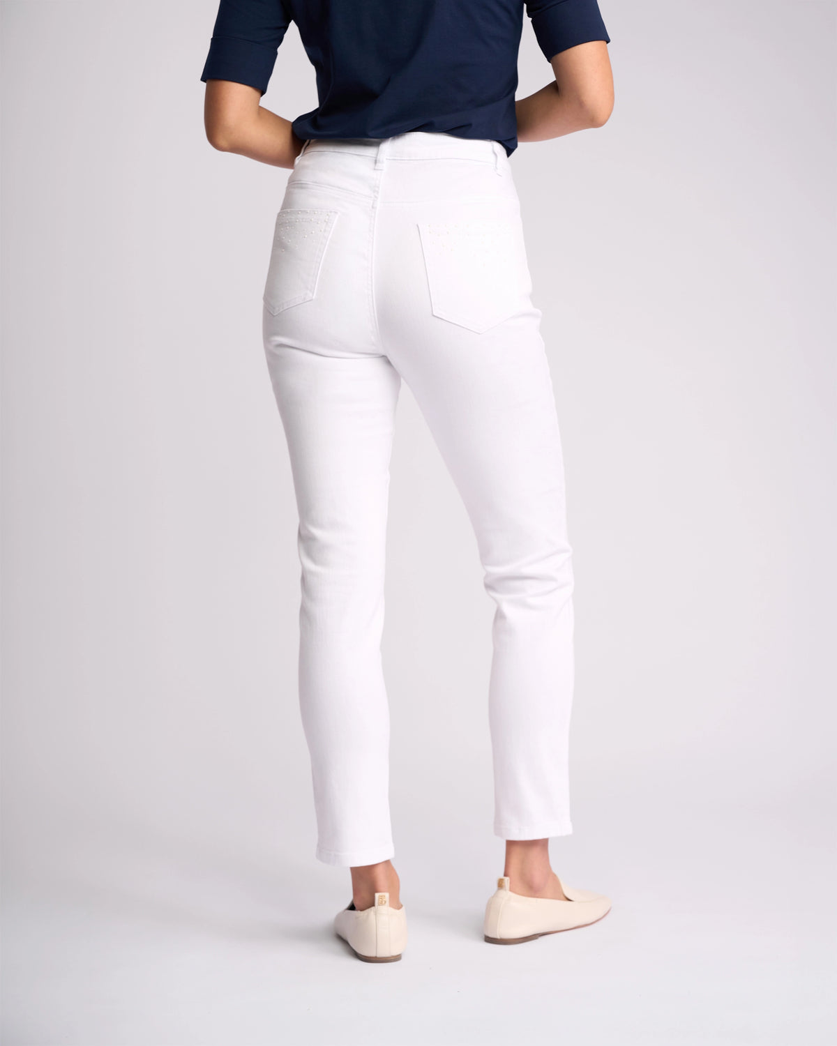 A person faces away, wearing the Vassalli Tide Jean in White Denim with a navy top and beige shoes, set against a plain light background.