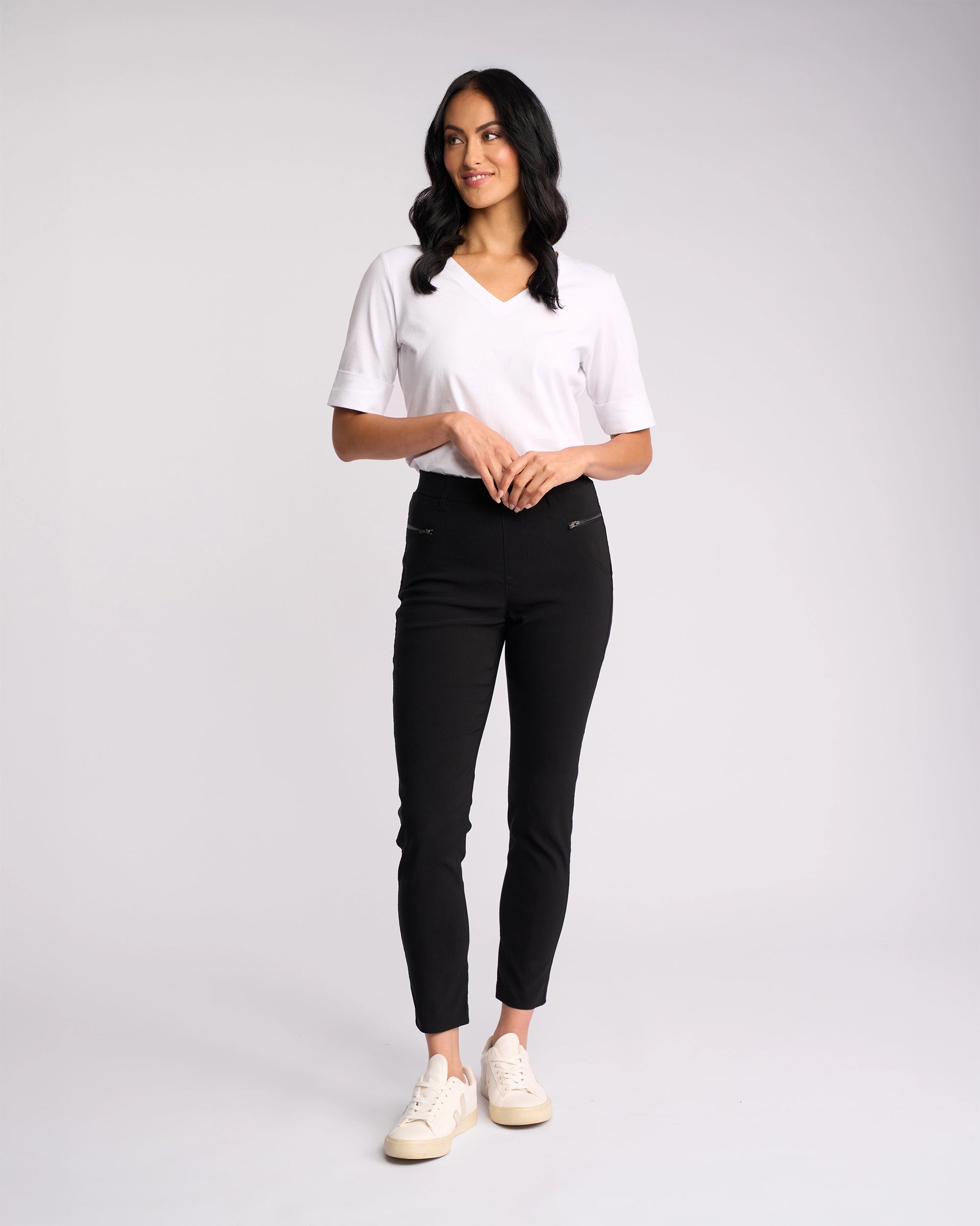 A woman with wavy, shoulder-length dark hair stands against a light background, wearing Vassalli's Concept Pant in black with a white short-sleeve V-neck shirt and white sneakers. She gazes slightly to the side in a relaxed pose.
