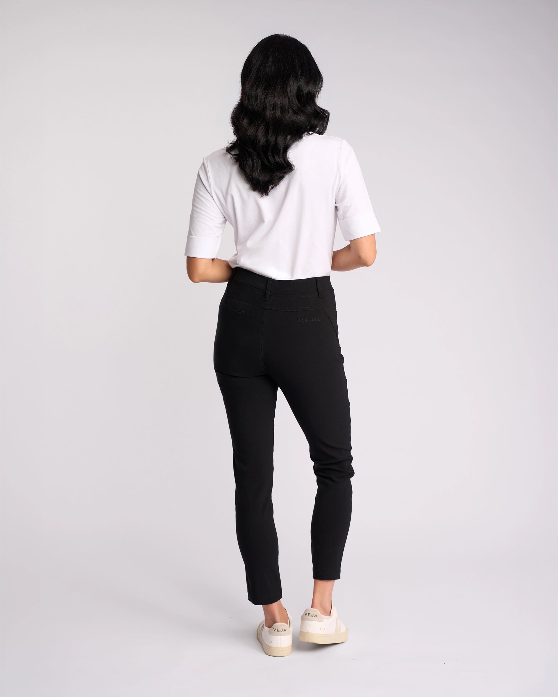 A person with long dark hair, wearing a white short-sleeve shirt, Vassalli Concept Pant - Black, and white sneakers stands facing away against a plain light background.