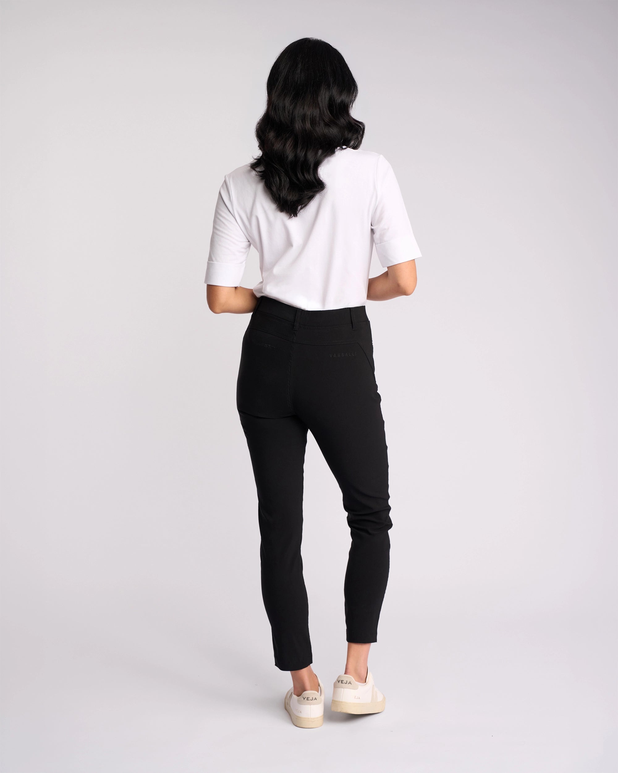 A person with long dark hair, wearing a white short-sleeve shirt, Vassalli Concept Pant - Black, and white sneakers stands facing away against a plain light background.
