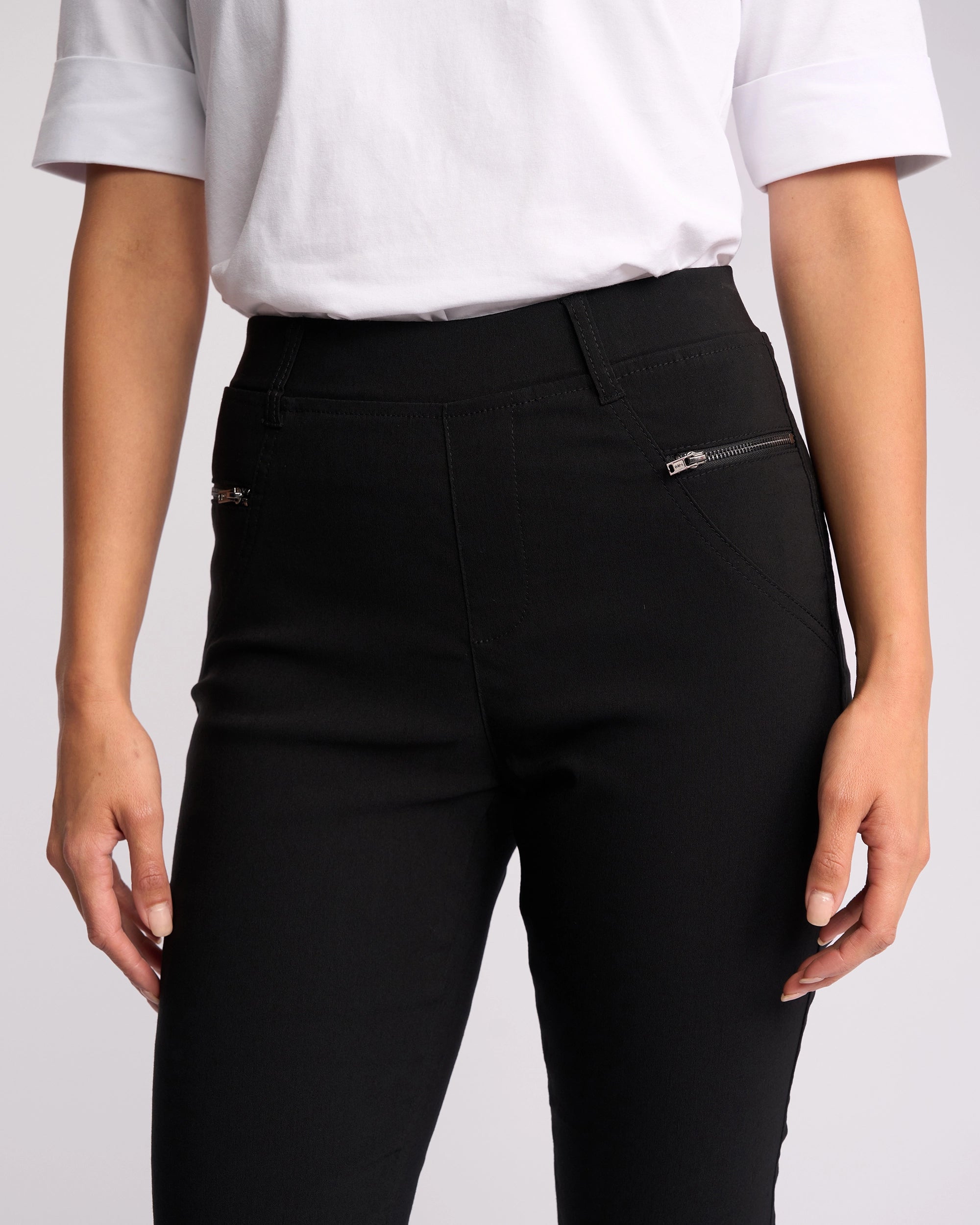 A person wearing a white short-sleeve top and Vassalli Concept Pant - Black, featuring high-rise fit with silver zippered pockets, shown from shoulders to mid-thigh.