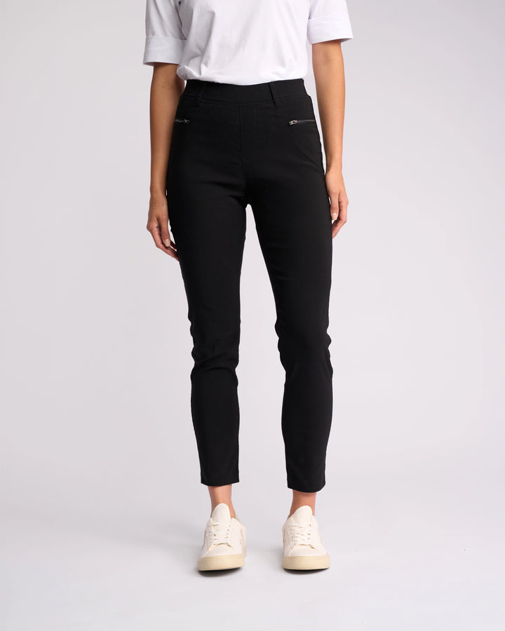A person models the Vassalli Concept Pant - Black, styled with a white short-sleeve shirt and white sneakers, standing against a plain light background. The model's head is not visible in the image.