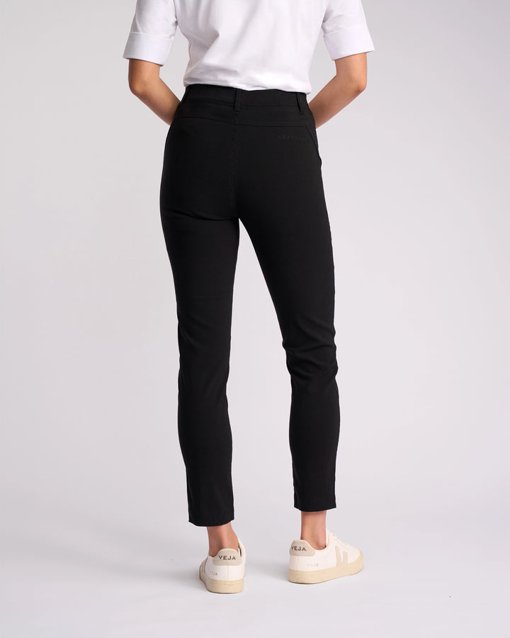 A person stands facing away against a plain light background, wearing a white short-sleeve shirt, Vassalli Concept Pant - Black (high-rise ankle-grazer), and white sneakers with beige soles.