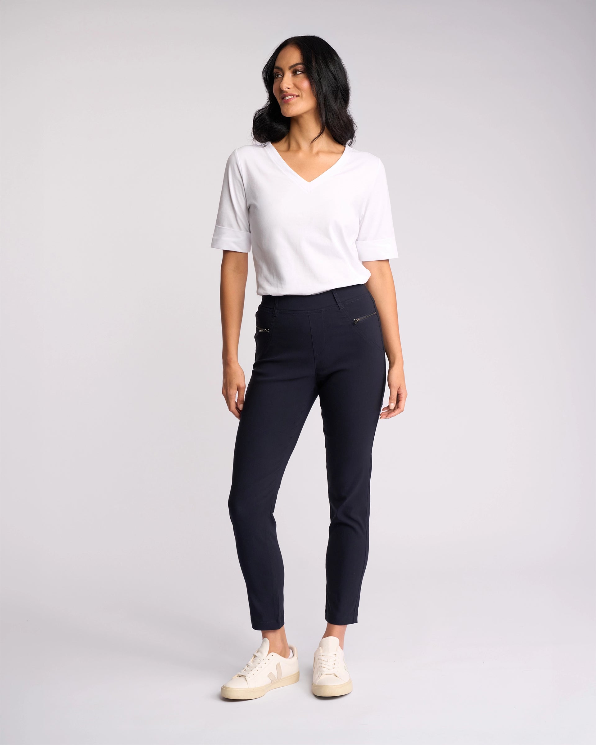 A woman stands against a plain light background, smiling as she looks to the side. She wears a white V-neck short-sleeve top, Vassalli Concept Pant - Navy with zipper details, and cream-colored sneakers.