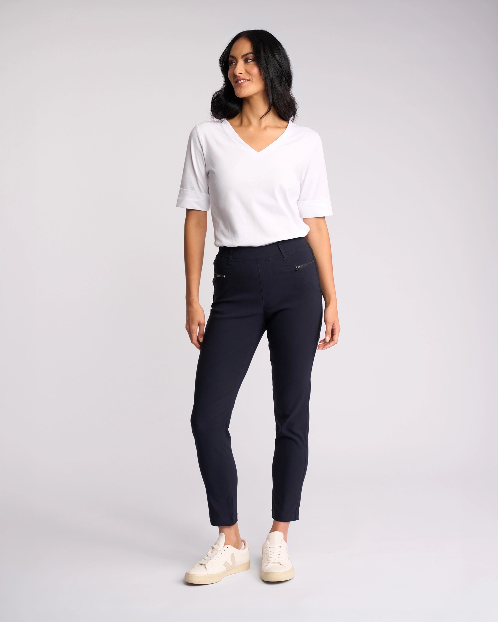 A woman stands against a plain light background, smiling as she looks to the side. She wears a white V-neck short-sleeve top, Vassalli Concept Pant - Navy with zipper details, and cream-colored sneakers.