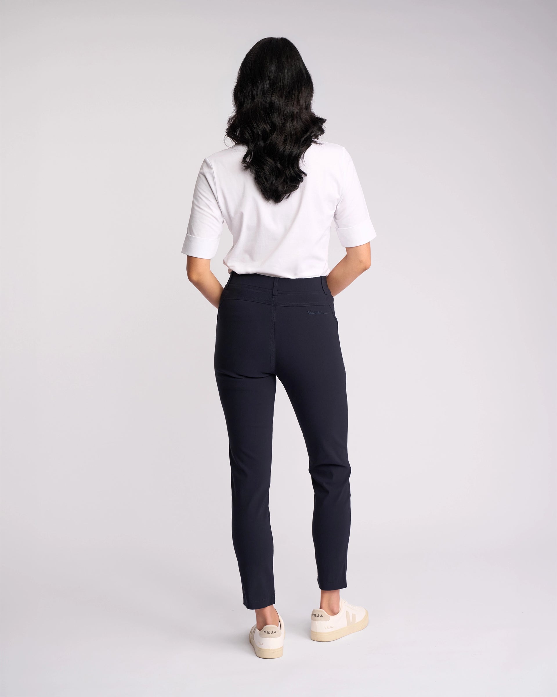 A person with long dark hair stands facing away, wearing a white short-sleeve shirt, white sneakers, and Vassalli’s Concept Pant - Navy against a plain light background.