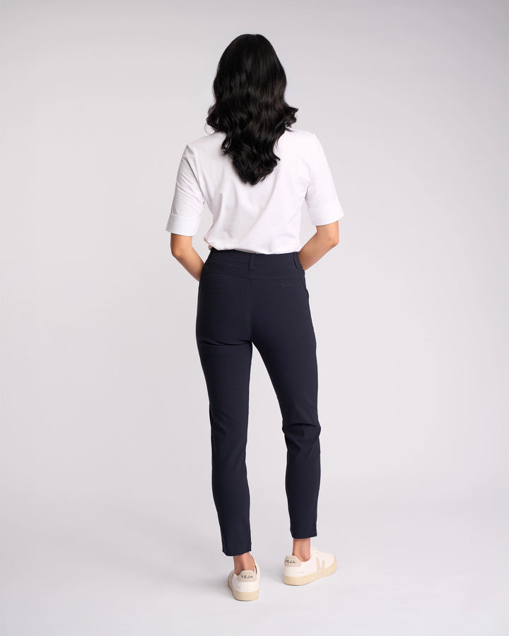 A person with long dark hair stands facing away, wearing a white short-sleeve shirt, white sneakers, and Vassalli’s Concept Pant - Navy against a plain light background.