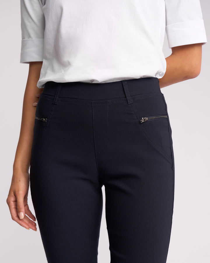 A person stands with one arm relaxed, wearing the Vassalli Concept Pant in navy—high-rise, skinny fit, made from stretch fabric and featuring two front zippered pockets—with a white short-sleeve shirt.