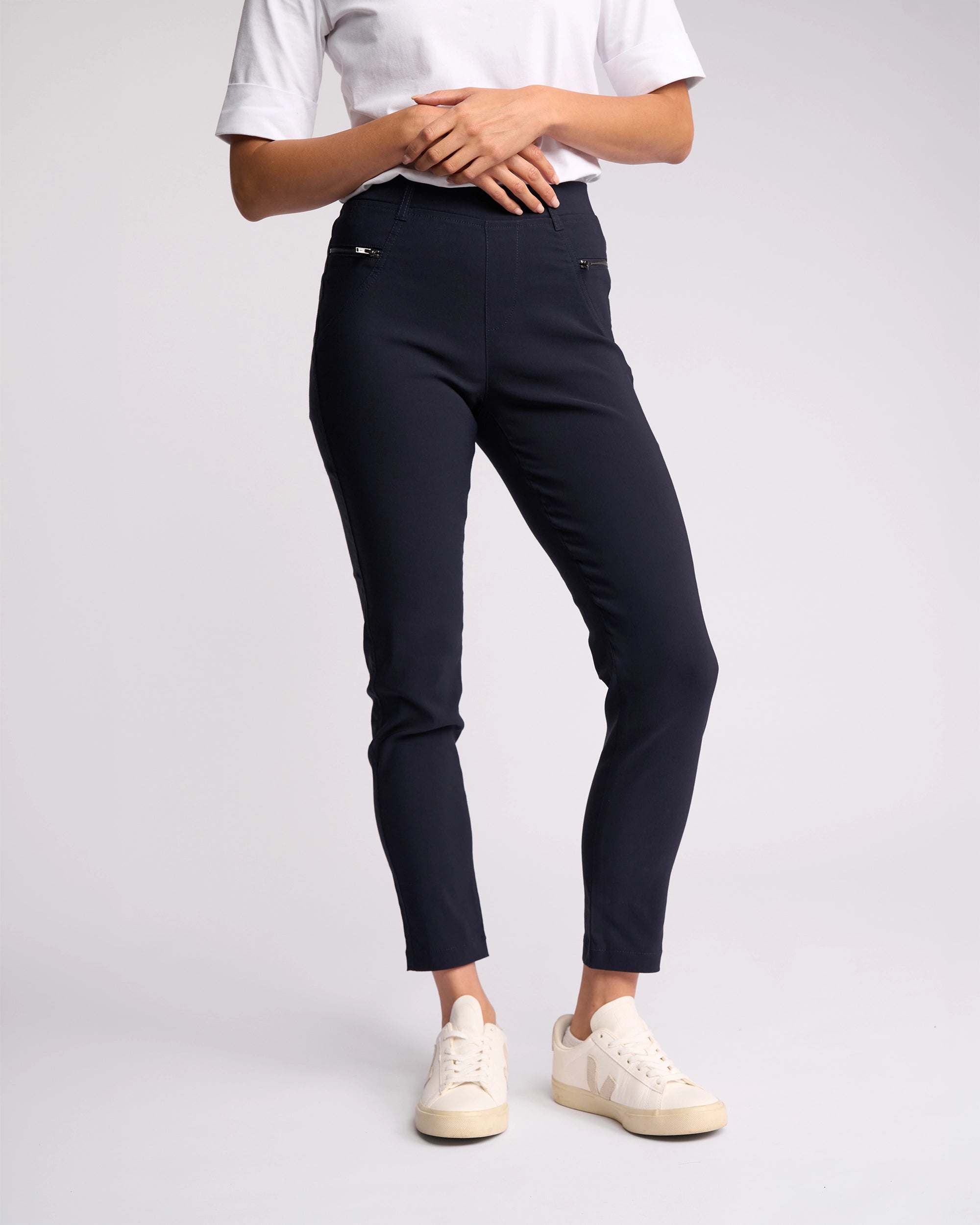 A person in a white short-sleeve top, Vassalli Concept Pant - Navy, and off-white sneakers stands against a plain light background with hands near the chest.