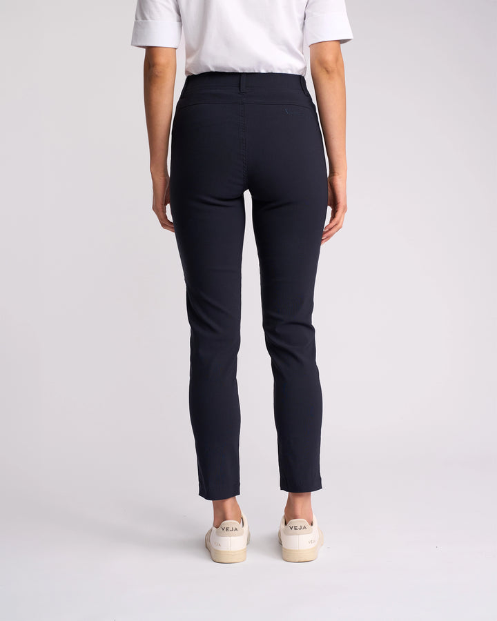 A person stands facing away from the camera, wearing a white short-sleeve shirt, Vassalli Concept Pant - Navy, and cream sneakers with VEJA branding on the back. The background is plain and light-colored.