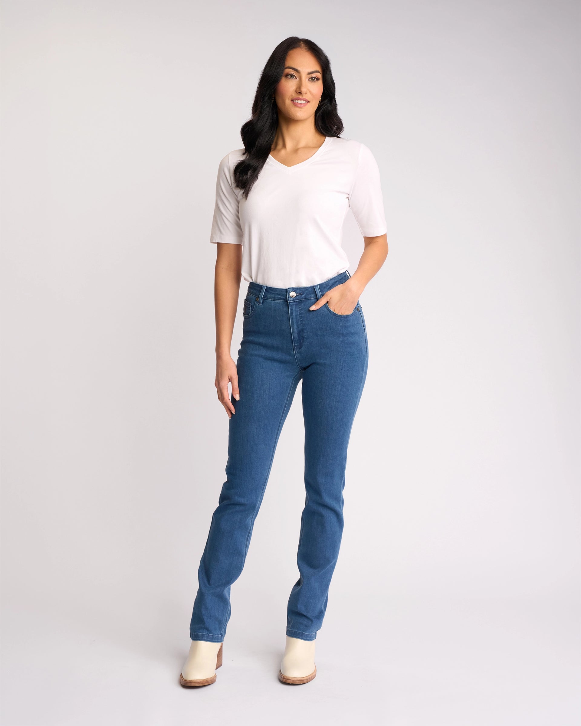 A woman with long dark hair stands against a plain background, smiling with one hand in her pocket. She wears a white V-neck shirt, light-colored shoes, and Vassalli’s Onset Jean - Plain New Blue high-rise slim-leg denim jeans.