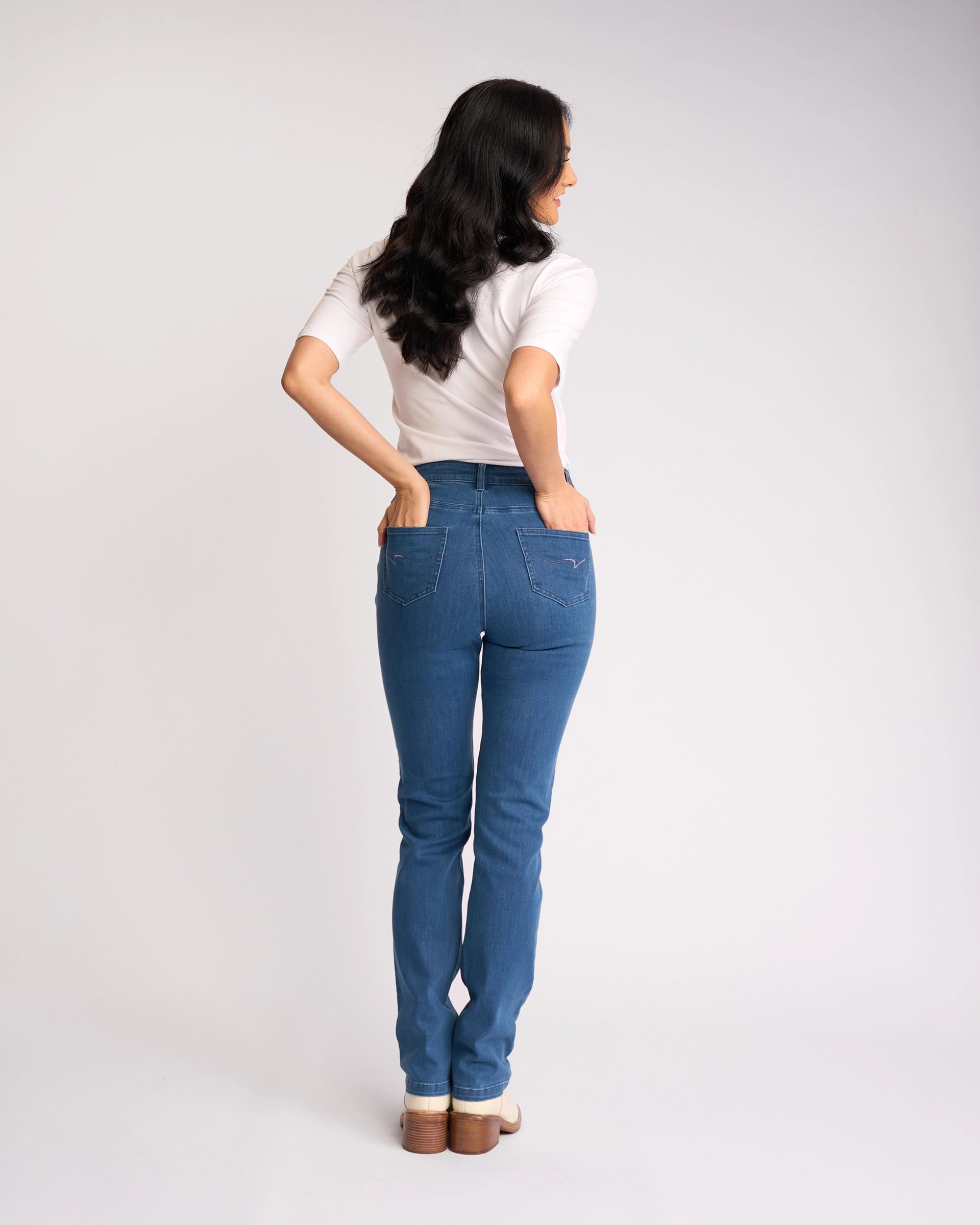 A woman with long dark hair, facing away, wears a white short-sleeve shirt, beige shoes, and Vassalli Onset Jean - Plain New Blue high-rise slim-leg stretch denim jeans against a plain white background.