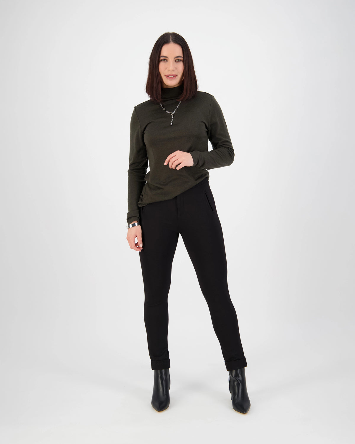 A woman with straight dark hair stands against a white background, wearing a dark green long-sleeve top, Vassalli Style Pant - Black, black ankle boots, and a necklace for an effortlessly polished workwear look as she smiles at the camera.