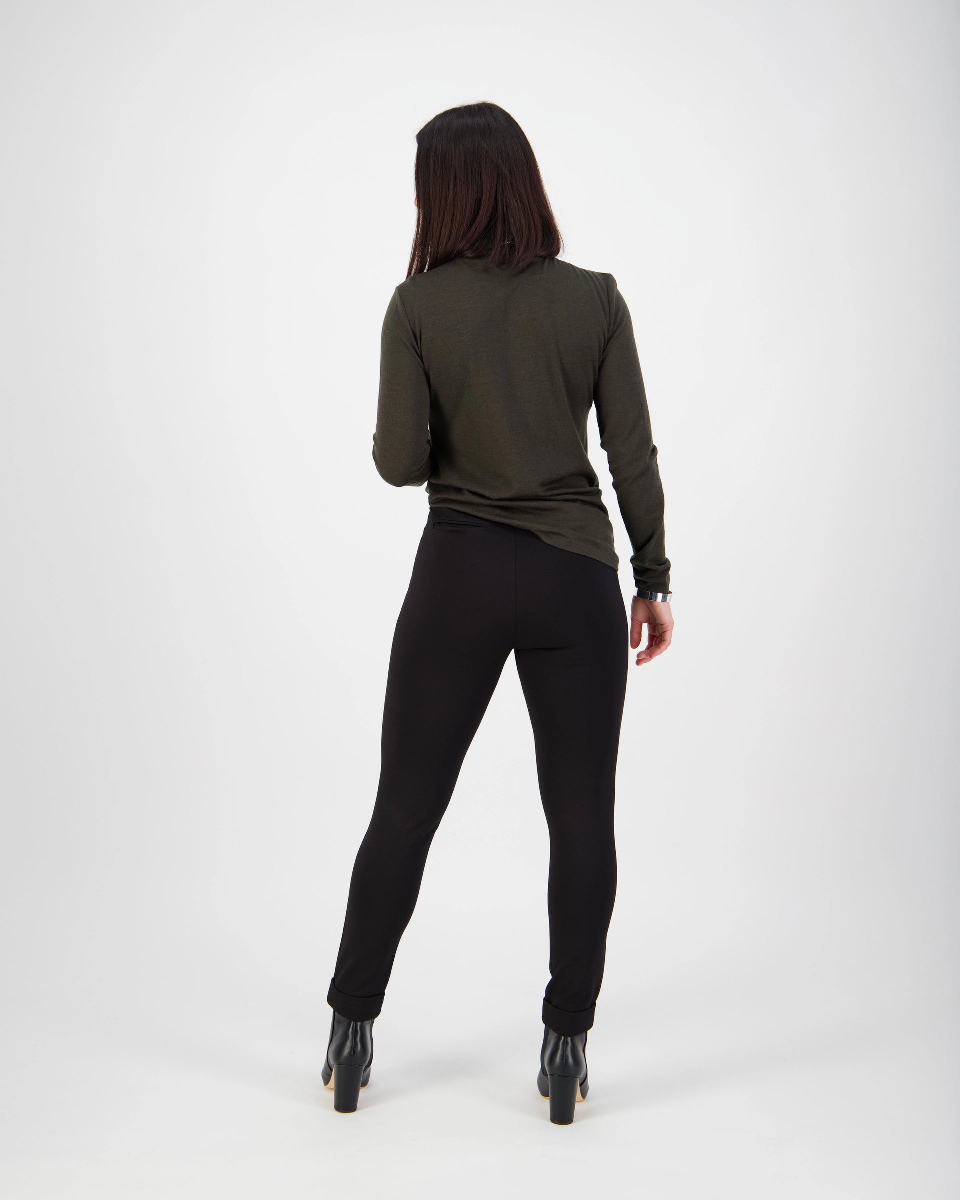 A person with long dark hair, wearing a dark green long-sleeve top and Vassalli Style Pant - Black, stands facing away from the camera against a plain white background.