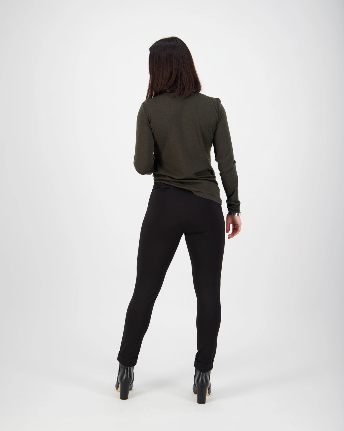 A person with long dark hair, wearing a dark green long-sleeve top and Vassalli Style Pant - Black, stands facing away from the camera against a plain white background.