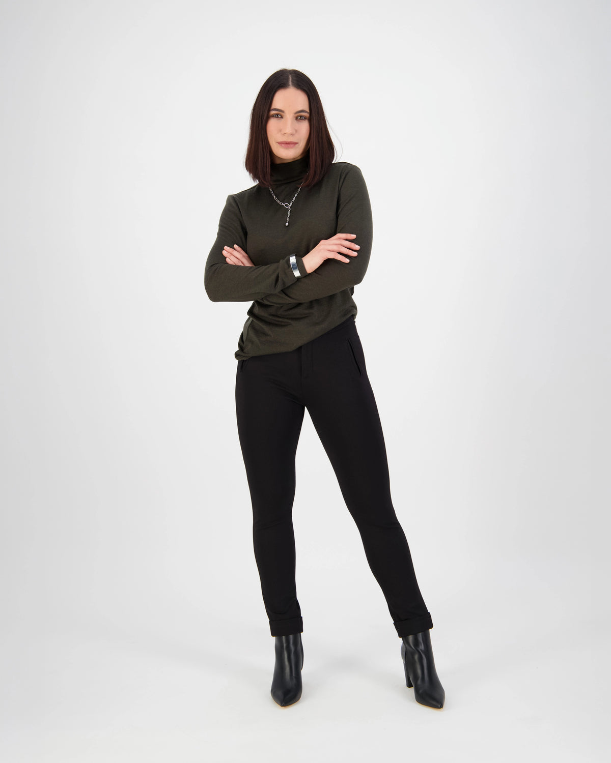 A woman with straight dark hair stands confidently with arms crossed, wearing a dark green long-sleeve top, Vassalli Style Pant - Black, and black ankle boots against a plain white background.