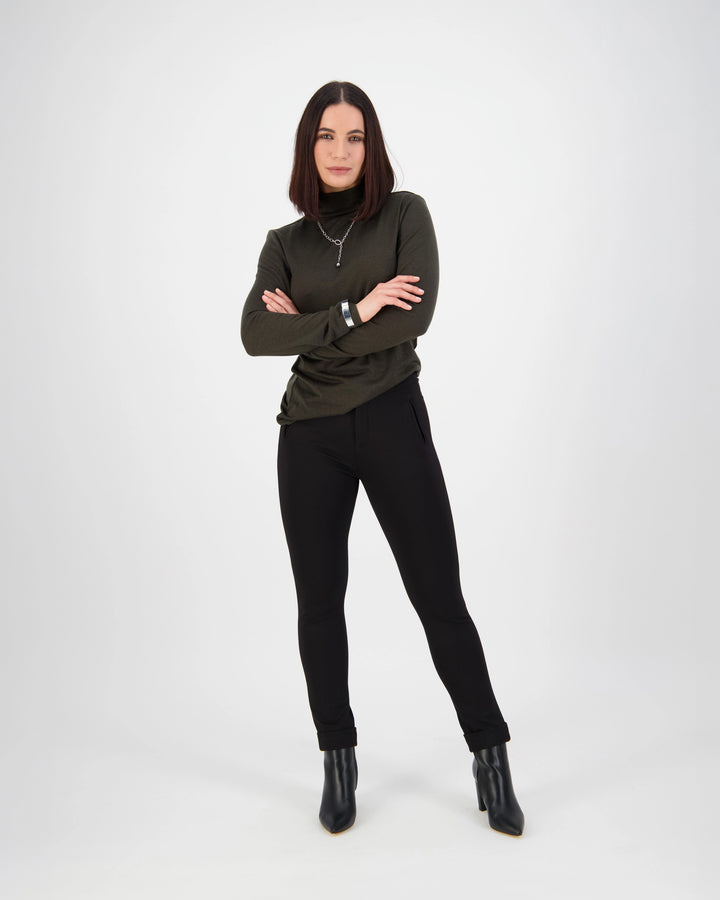 A woman with straight dark hair stands confidently with arms crossed, wearing a dark green long-sleeve top, Vassalli Style Pant - Black, and black ankle boots against a plain white background.