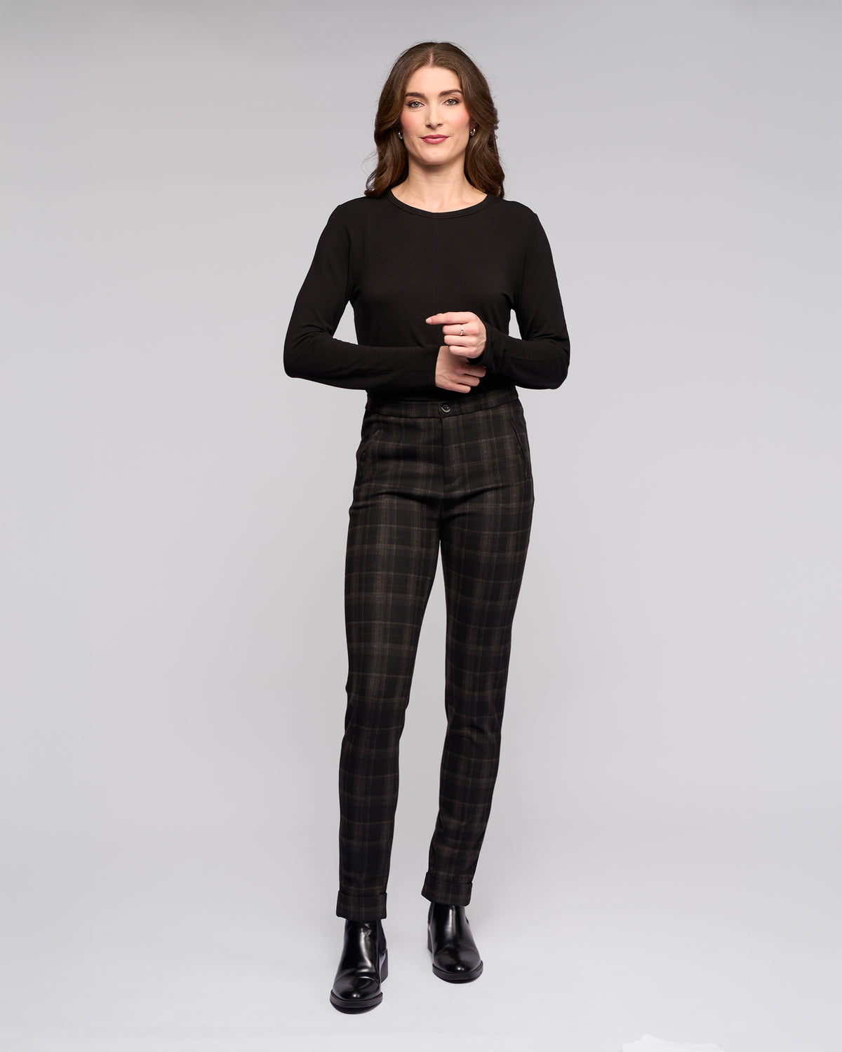A woman with long brown hair smiles softly, standing against a light gray background in a black long-sleeve top and Vassalli Style Pant - Checkmate, paired with black ankle boots and her hands clasped gently.
